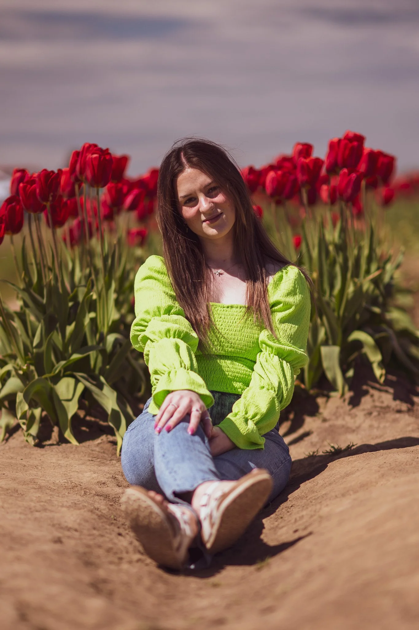 A young woman sitting on the ground in front of a row of red tulips, wearing a bright green top and jeans.