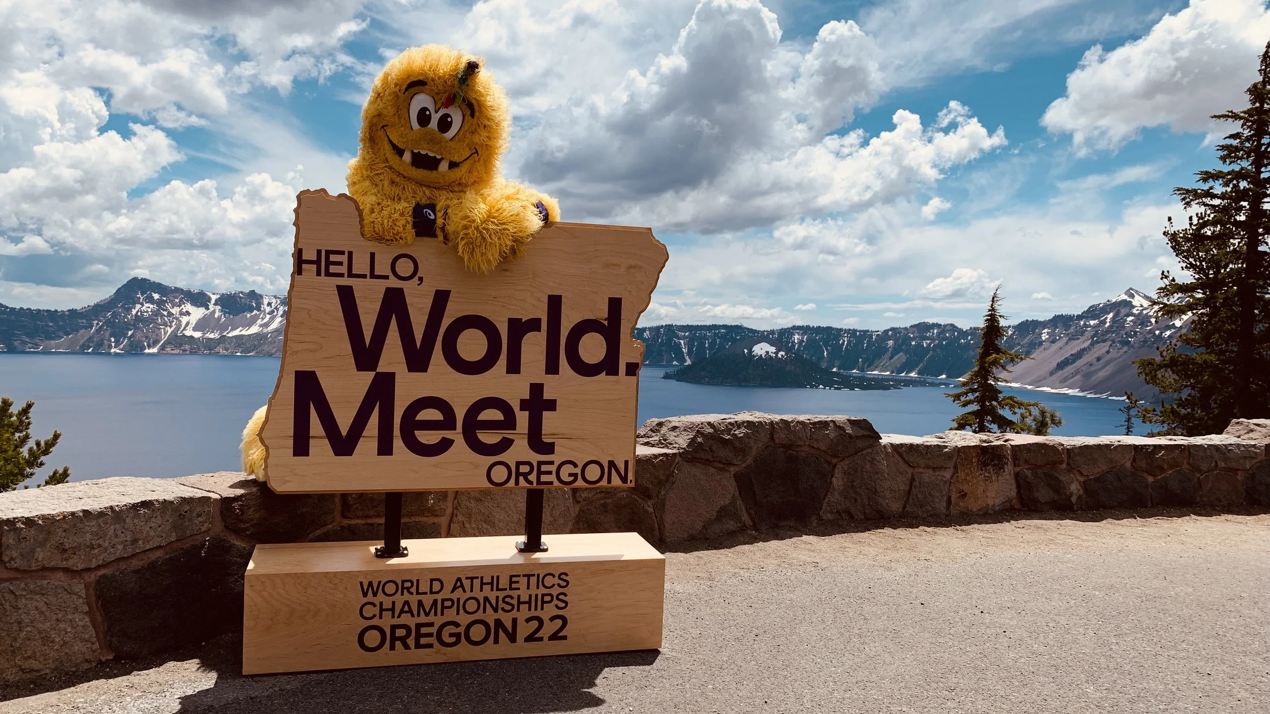 A mascot with yellow fur and a big smile standing behind a sign that reads "Hello, World. Meet Oregon" near a lake with mountains in the background.