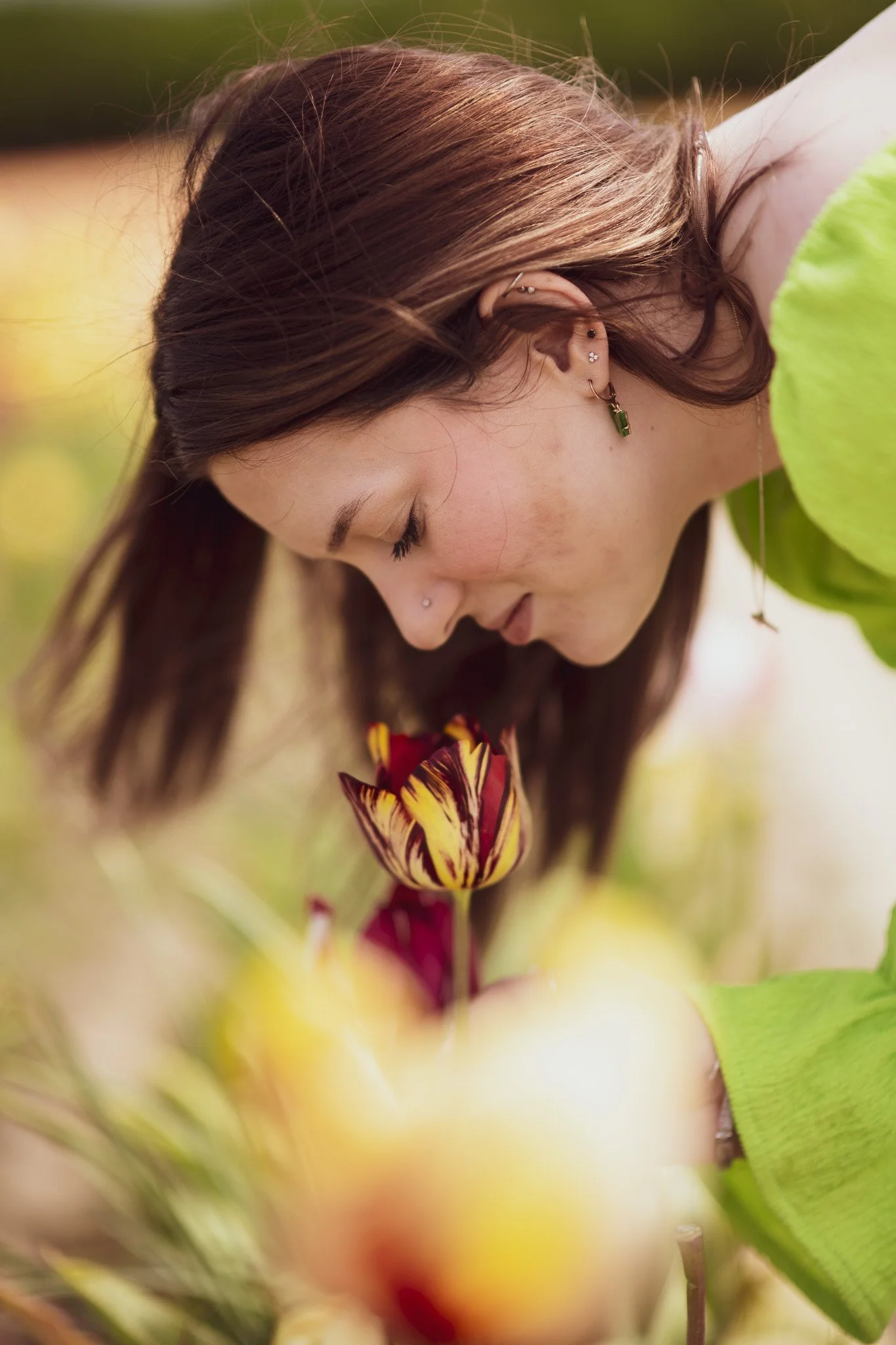 A young woman with brown hair and multiple earrings is smelling a yellow and red tulip in a garden, with blurred yellow flowers in the background.