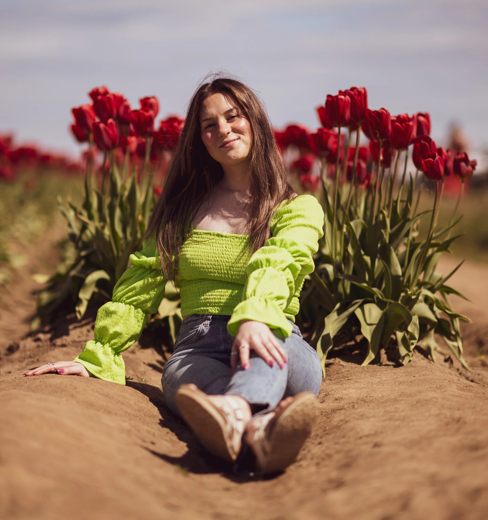 A woman sitting on the ground outdoors near a row of red tulips, wearing a bright green top and jeans, with a slight smile, during daytime.