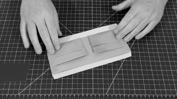 Hands assembling a paper model with folded paper strips on a cutting mat.
