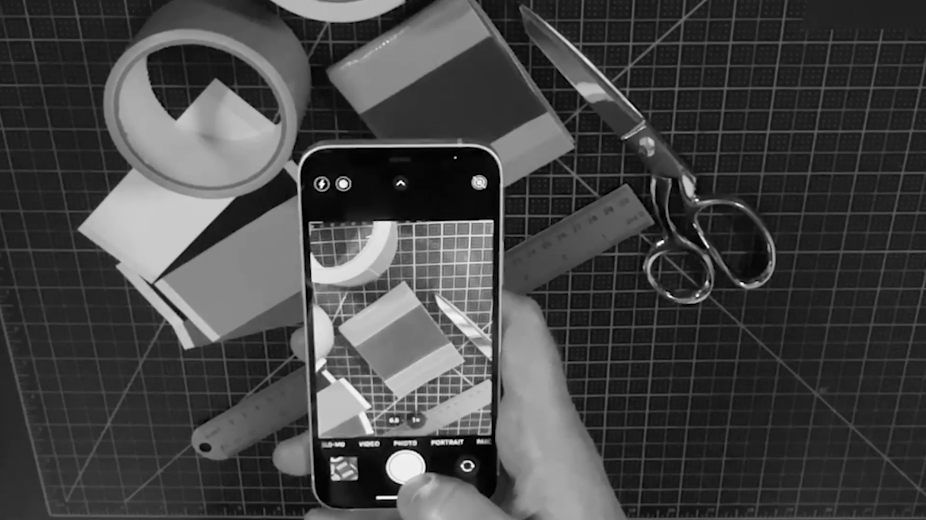A person is taking a photo of a workspace with scissors, tape, a ruler, and paper using a smartphone.