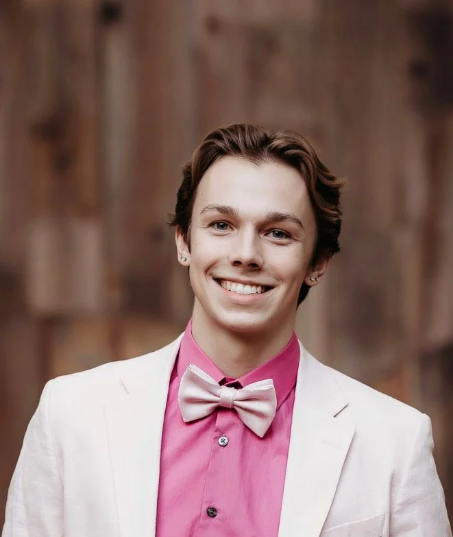Young man smiling, wearing a white suit jacket, pink dress shirt, and light pink bow tie, with a wooden wall background.