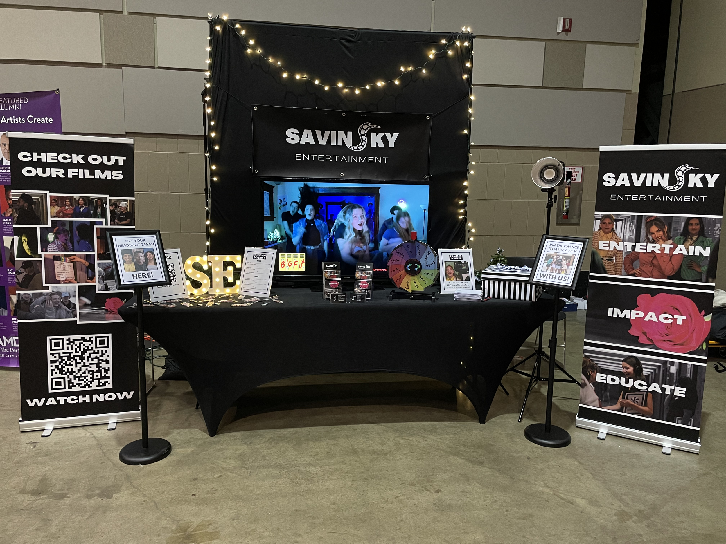 Booth for Savinsky Entertainment featuring a TV screen, promotional banners, a mini wheel, and informational signs, decorated with string lights.