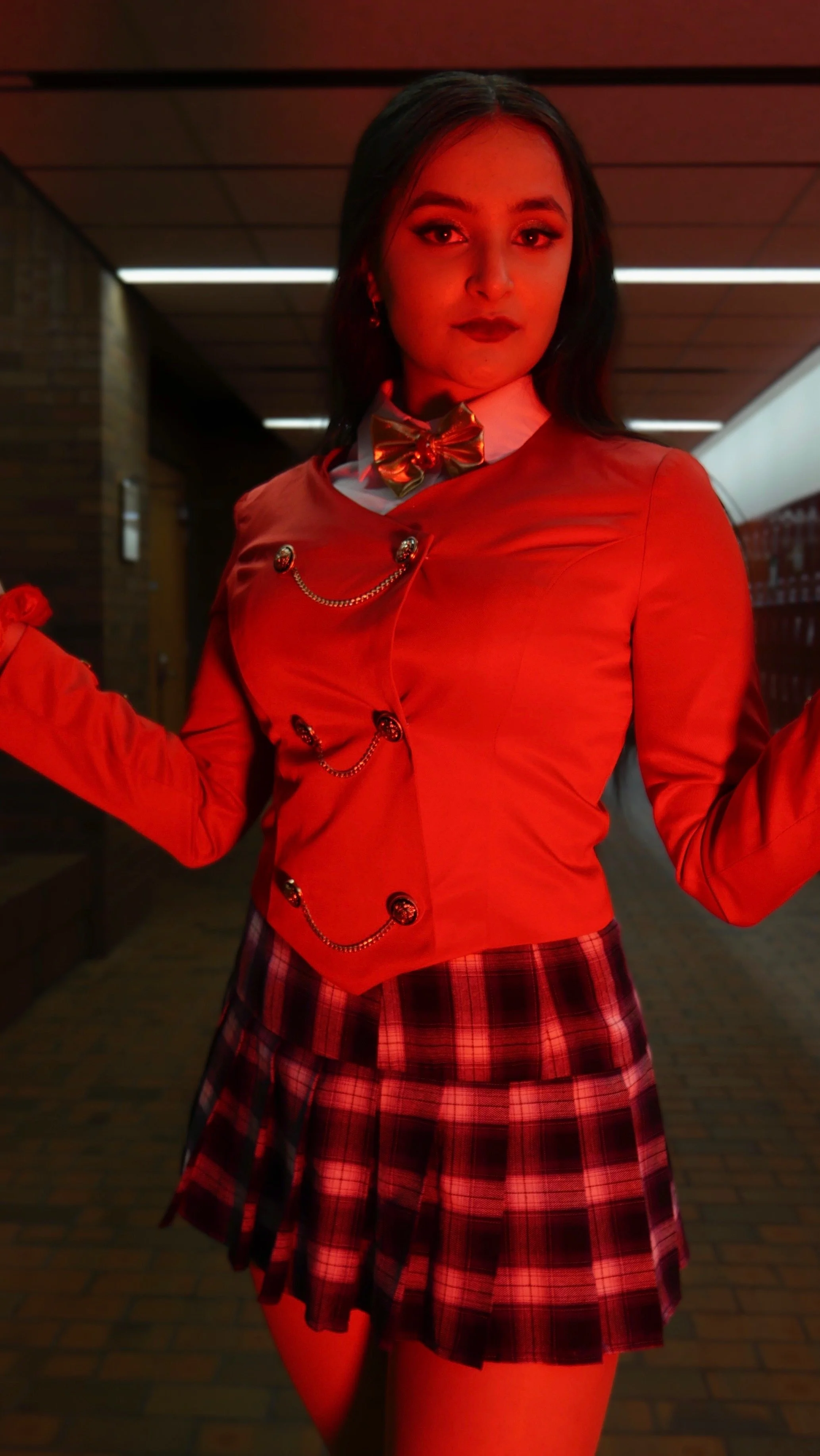 Young woman in a red blazer with decorative chains, white shirt with a gold bow tie, and a red plaid skirt, standing in a hallway with lockers.