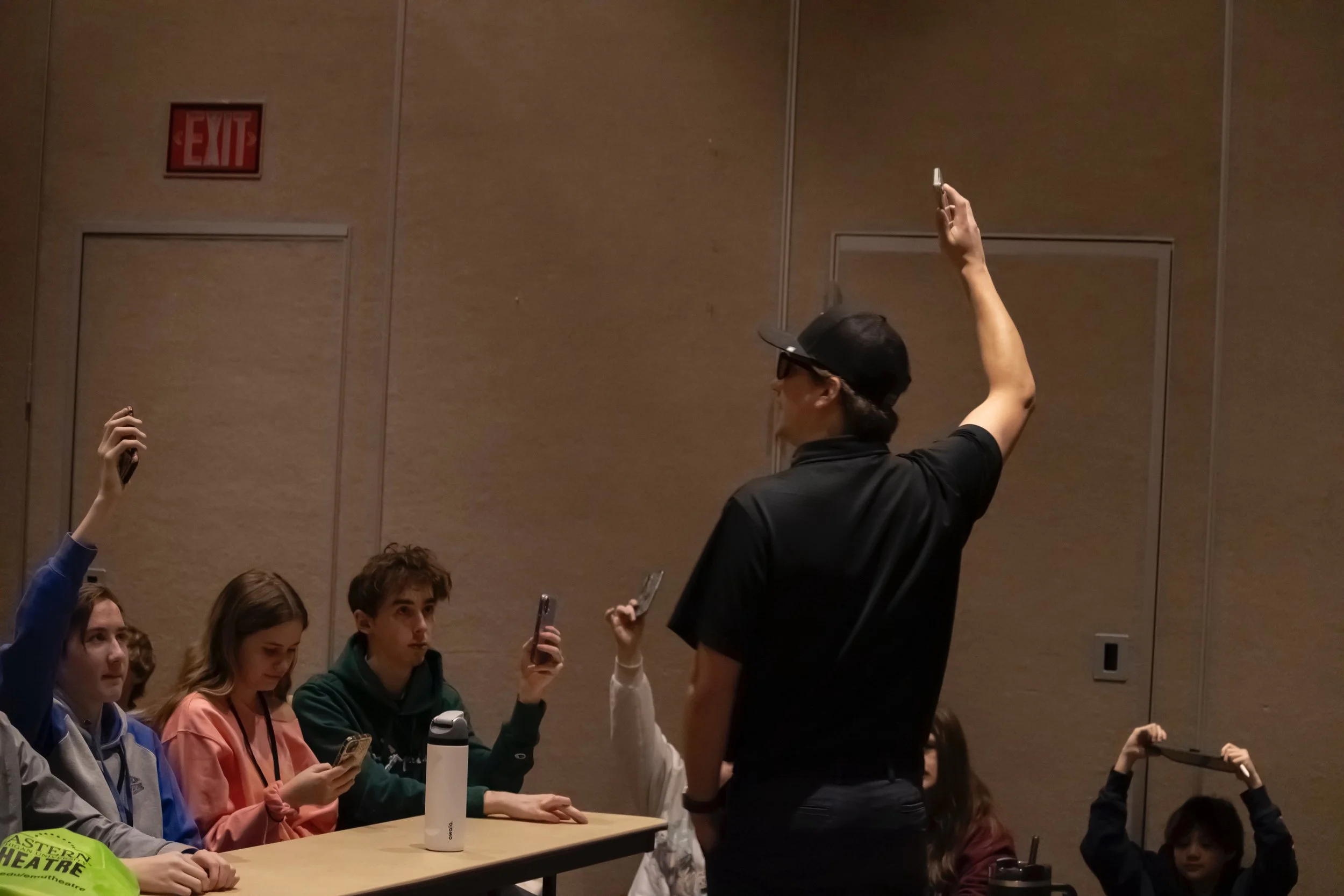 A group of students sitting at a table, raising hands, and taking photos or videos of a man standing in front of them, with one person holding a phone up to record or take a picture.