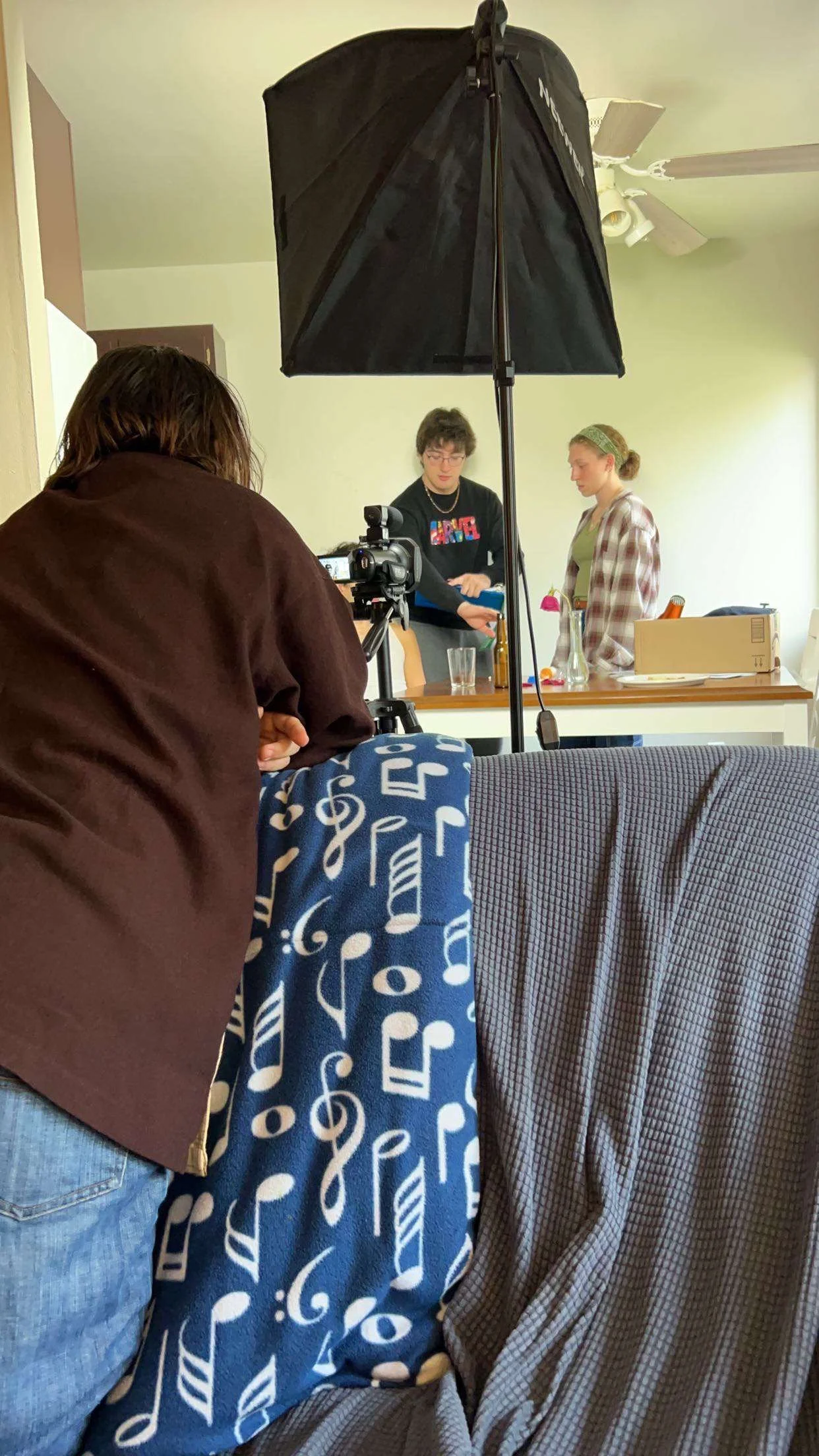 People filming a home video or recording in a living room, with a woman operating a camera on a tripod, a large softbox light overhead, and two individuals standing at a table in the background.