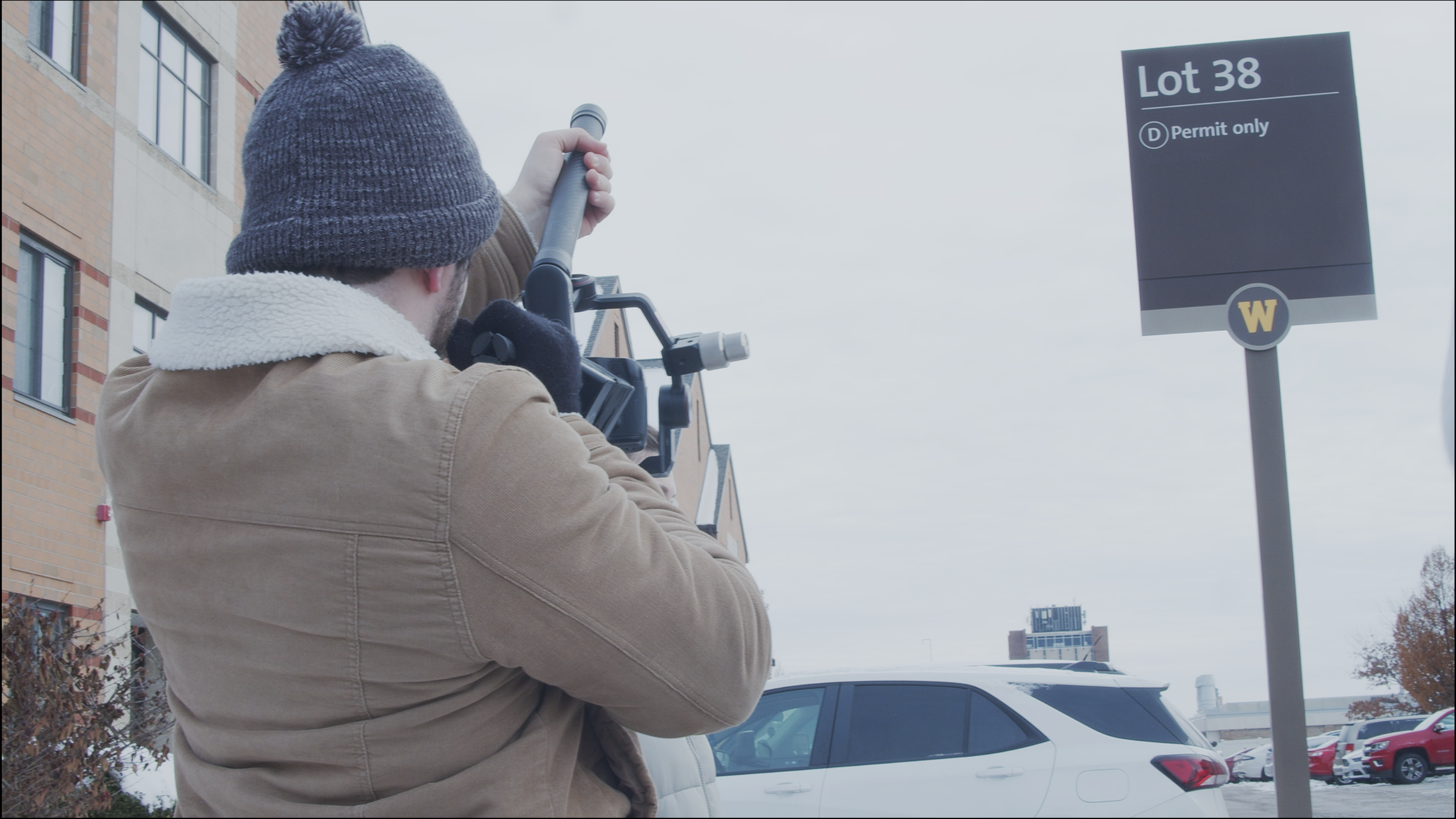 A person wearing a dark knit hat with a pom-pom, multitonal brown jacket with a white fleece collar, and black gloves is venturing a telescope in a parking lot with parked cars, including a white SUV. It is cloudy with an overcast sky. There is a sign indicating "Lot 38, Permit only" with a "W" emblem.