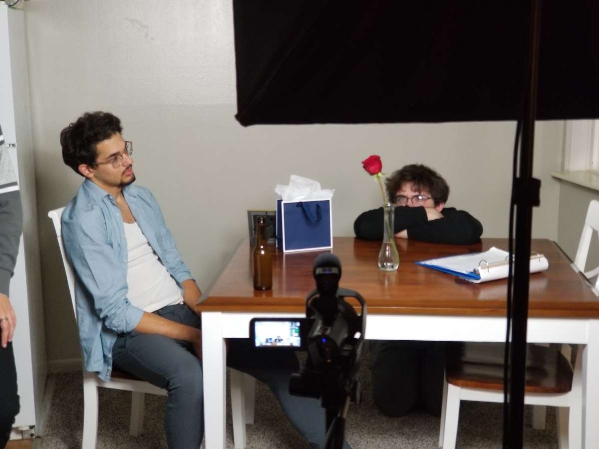 Two young men in a room with a wooden table. One is sitting in front of a camera, looking serious. The other is resting his head on the table with a rose in a glass in front of him, smiling. There are a gift bag, a notebook, and a brown bottle on the table. A studio light is visible in the foreground.