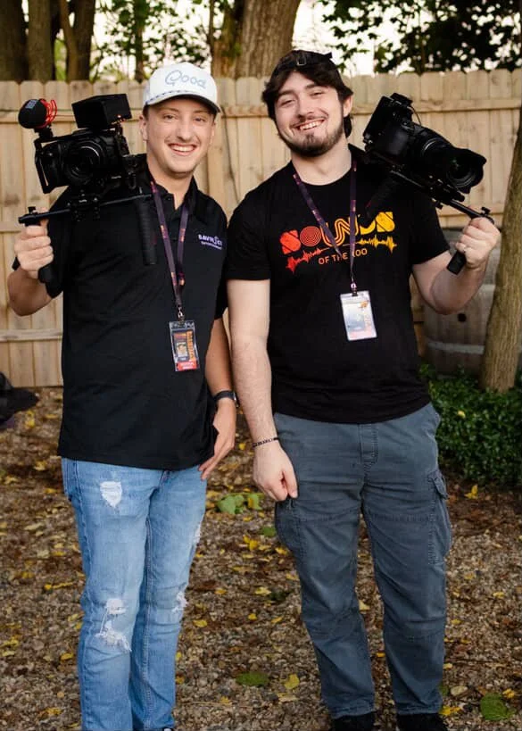 Two young men standing outdoors in front of a wooden fence, smiling, holding professional video cameras on their shoulders, wearing casual clothing with event badges around their necks.
