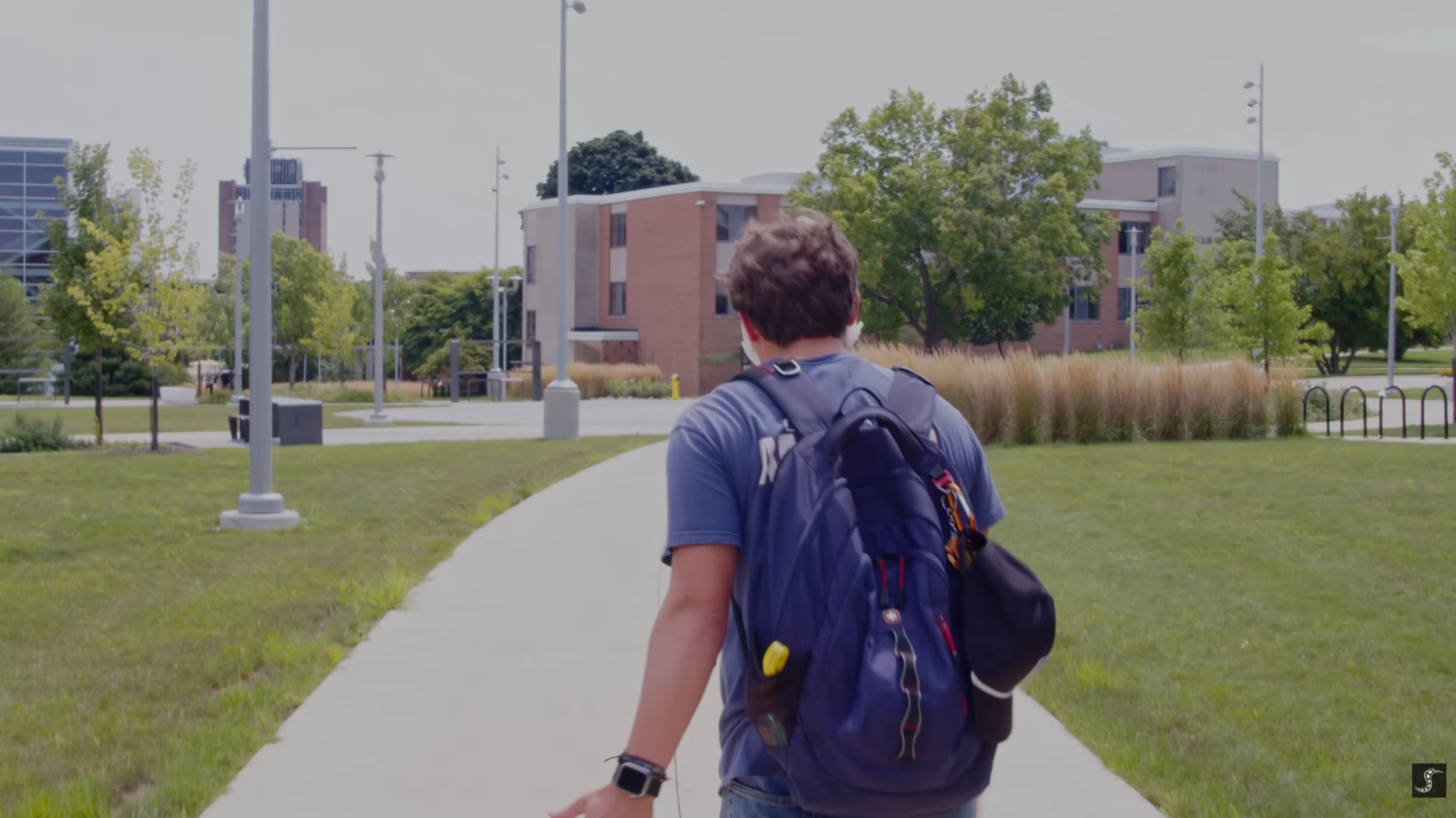 A young person with dark hair wearing a blue t-shirt and carrying a large navy backpack walks on a campus or park with green grass, trees, and modern buildings in the background.