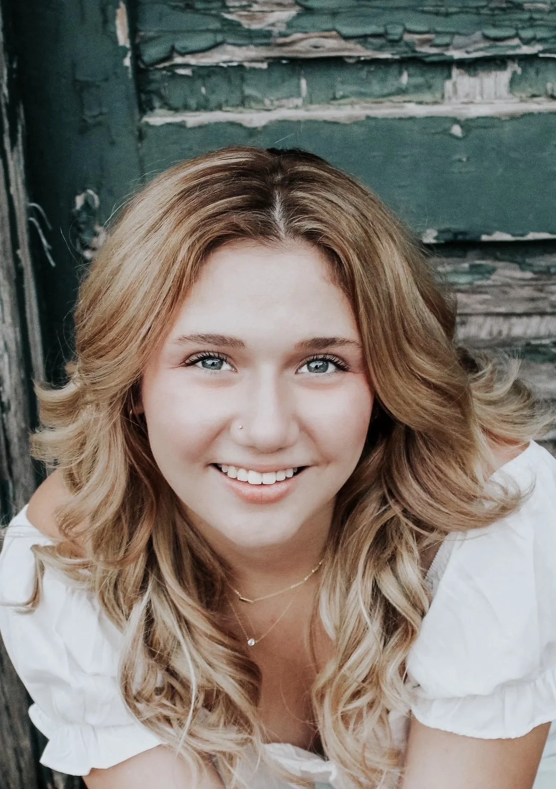A young woman with wavy, light brown hair and blue eyes smiling at the camera, wearing a white blouse and a gold necklace, with a weathered green wooden wall in the background.