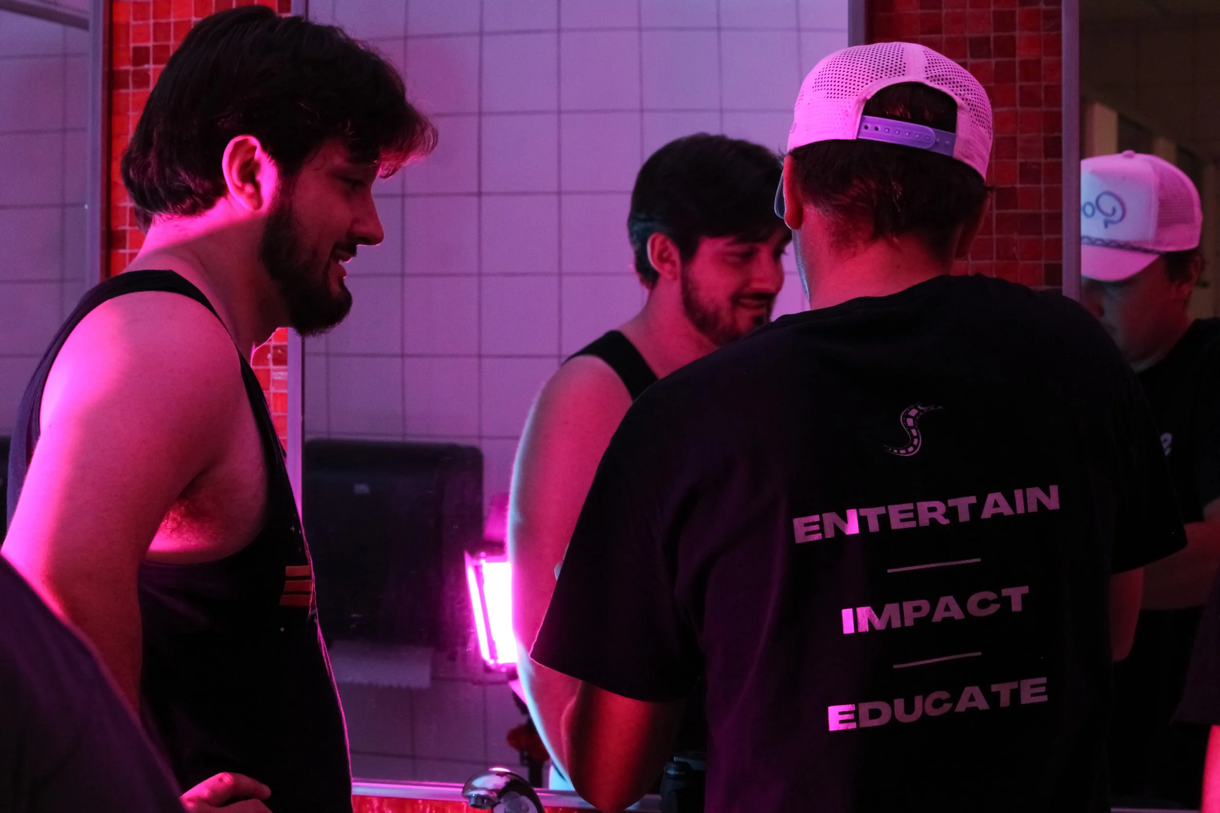 Four young men at an indoor event or party, talking and smiling under pink and purple lighting. Three are wearing black tank tops and one has a black shirt with words on the back, including "ENTERTAIN," "IMPACT," and "EDUCATE."