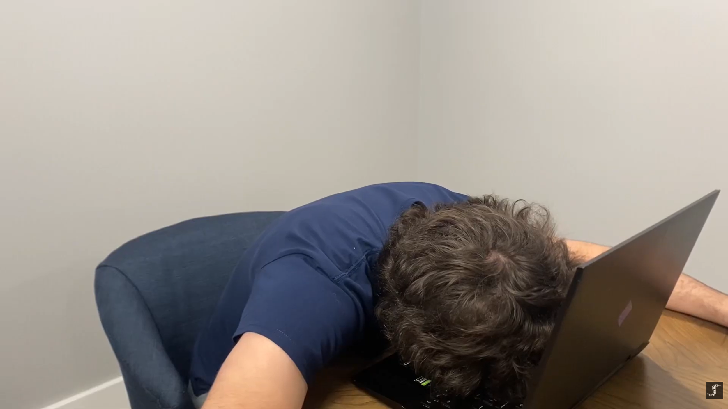 Person with curly hair wearing a navy t-shirt resting head on a laptop at a wooden table in a plain room.