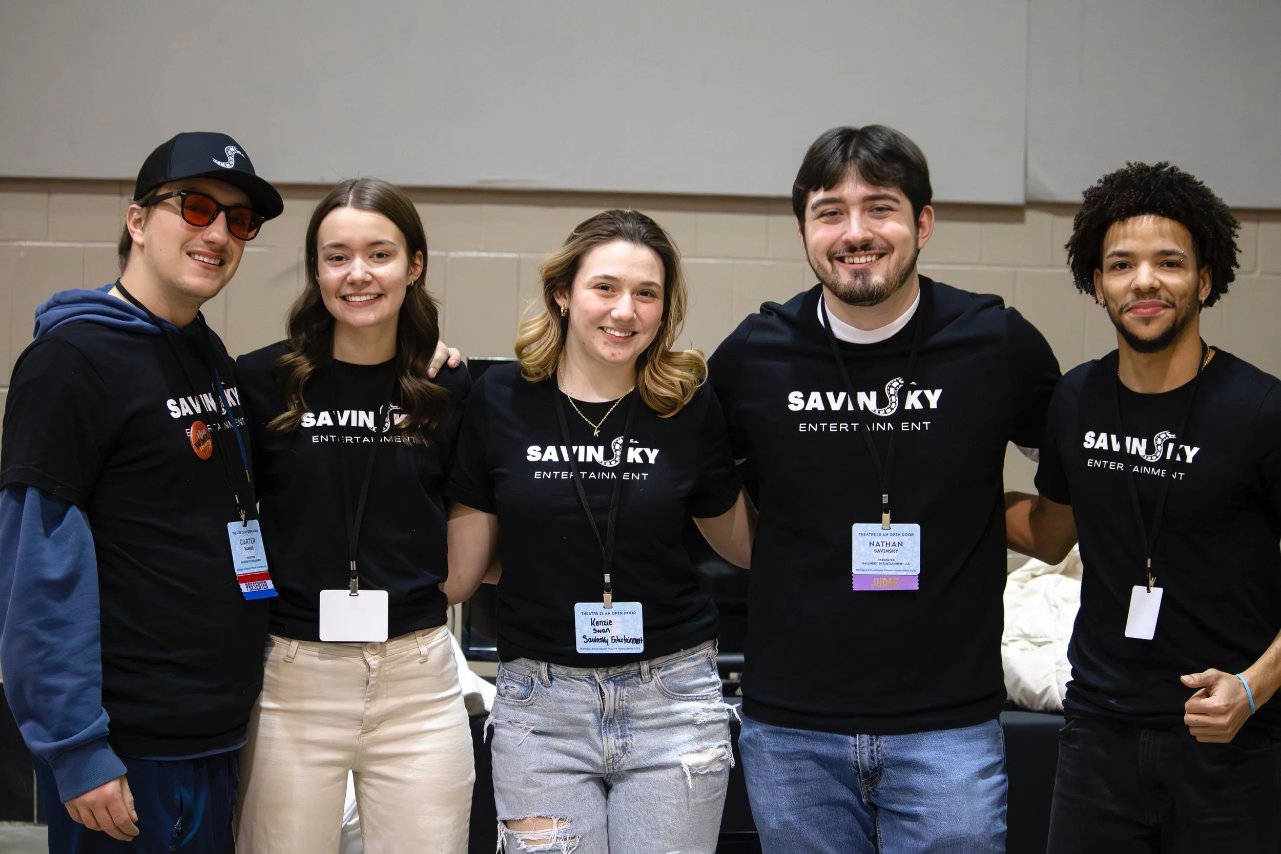 Group of five young people smiling and standing together, wearing matching black T-shirts with 'SAVIN SKY ENTERTAINMENT' printed on them, in an indoor setting.