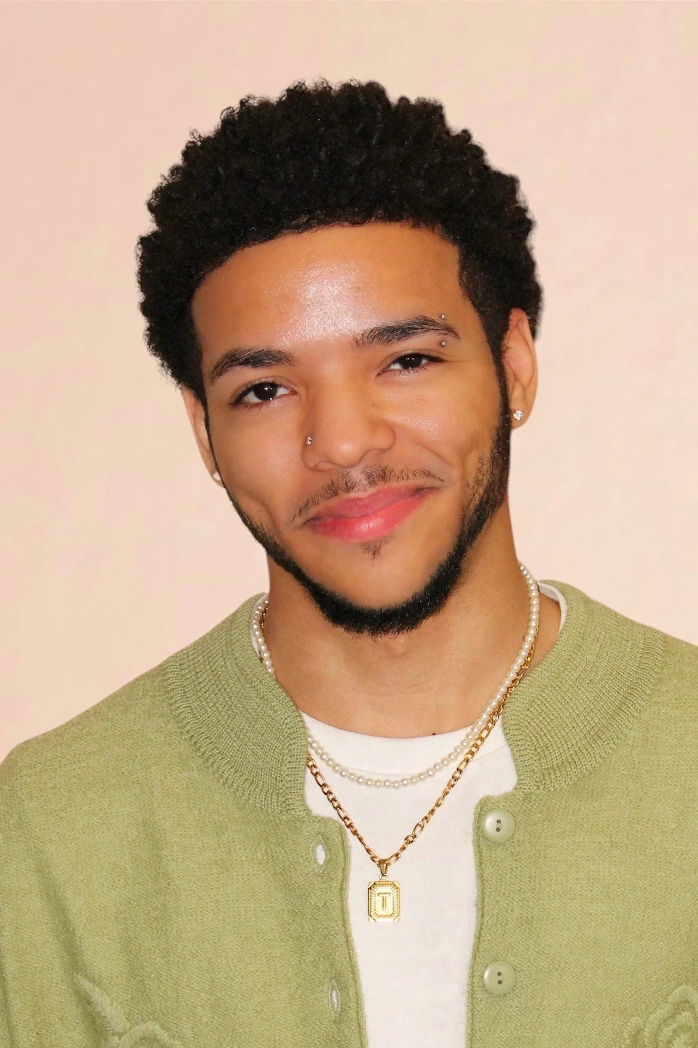 A young man with short curly hair, wearing earrings, a nose piercing, layered necklaces, and a light green cardigan, smiling and winking at the camera.