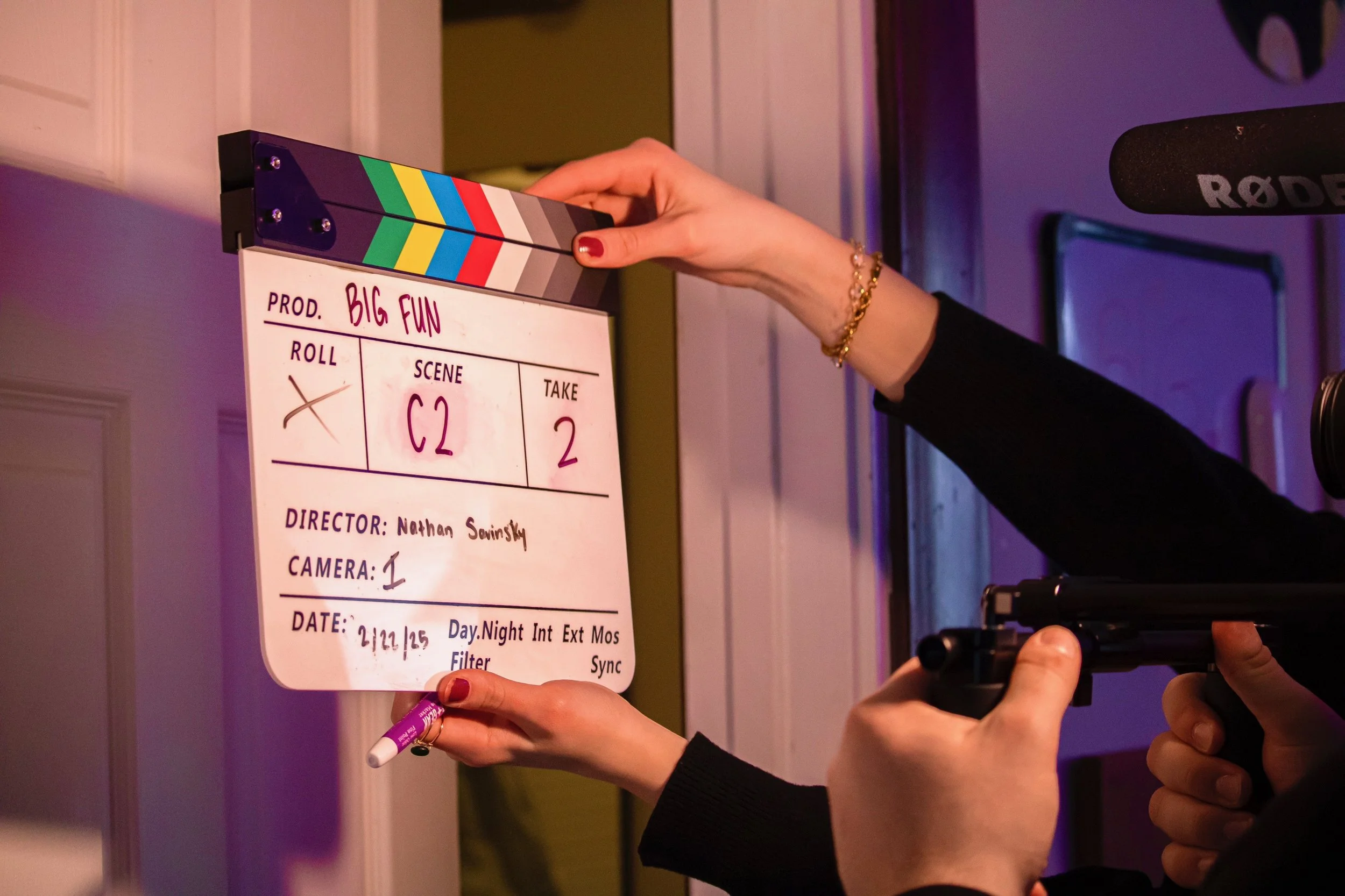 Person holding a film slate with a clapperboard, positioned near a film camera and microphone, in a film production setting.