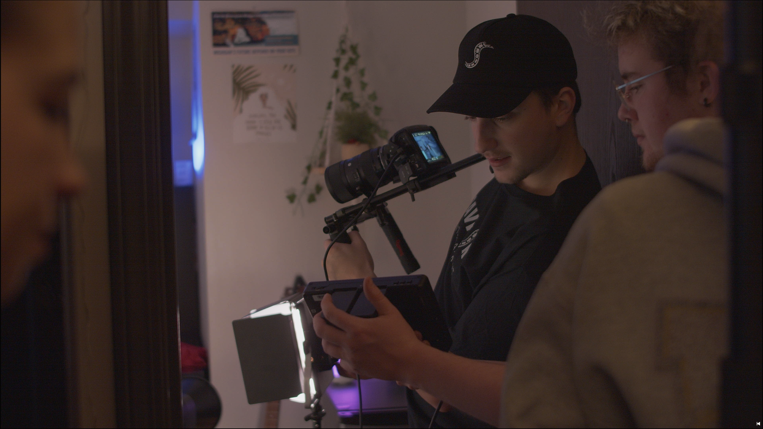 A person holding a video camera on a stabilizer while looking at a tablet device, with two other people nearby, in a room with a small plant and pictures on the wall.