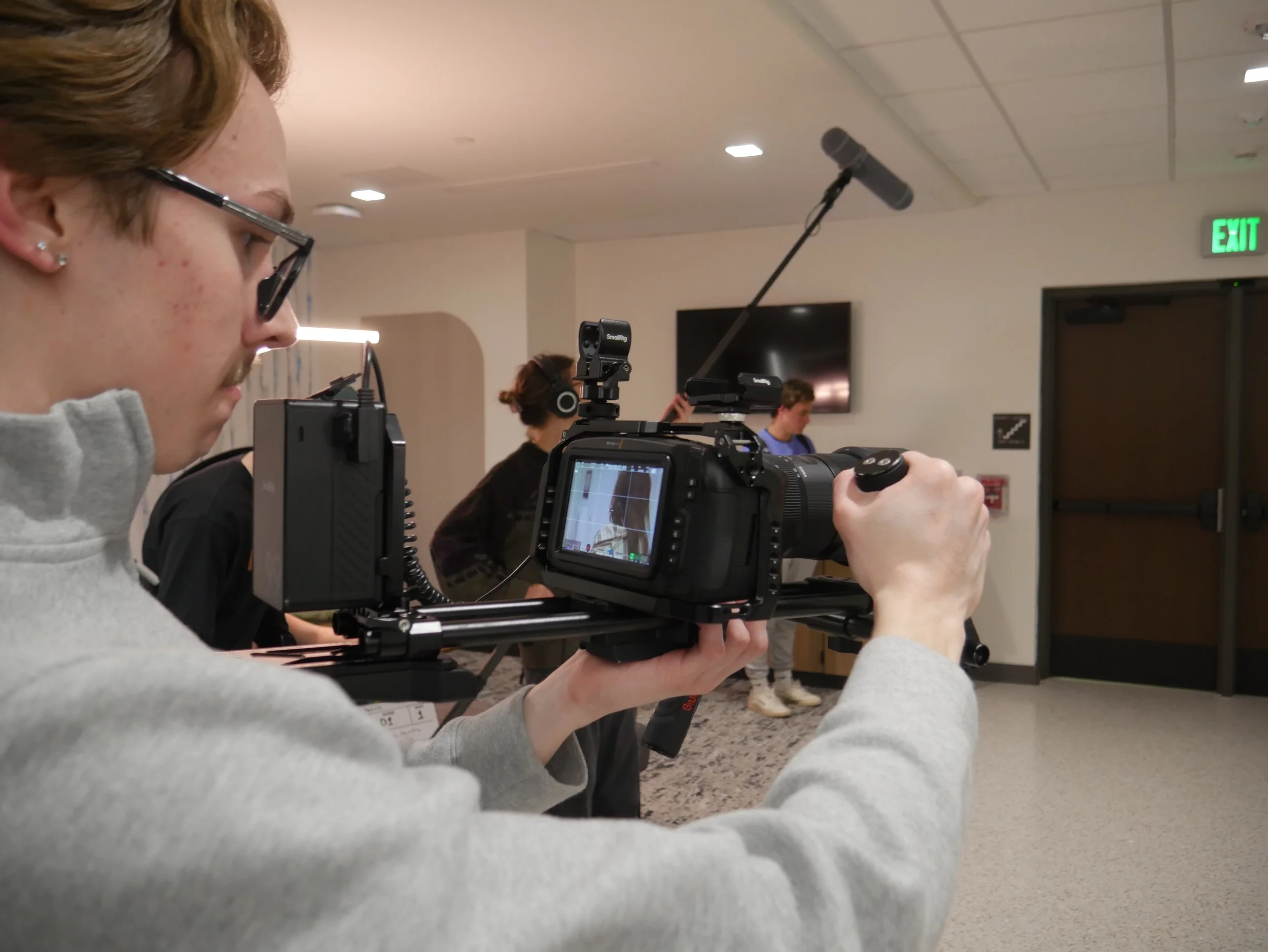 Person operating a professional camera with stabilizer rig in an indoor setting, likely filming or recording, with other individuals in the background.