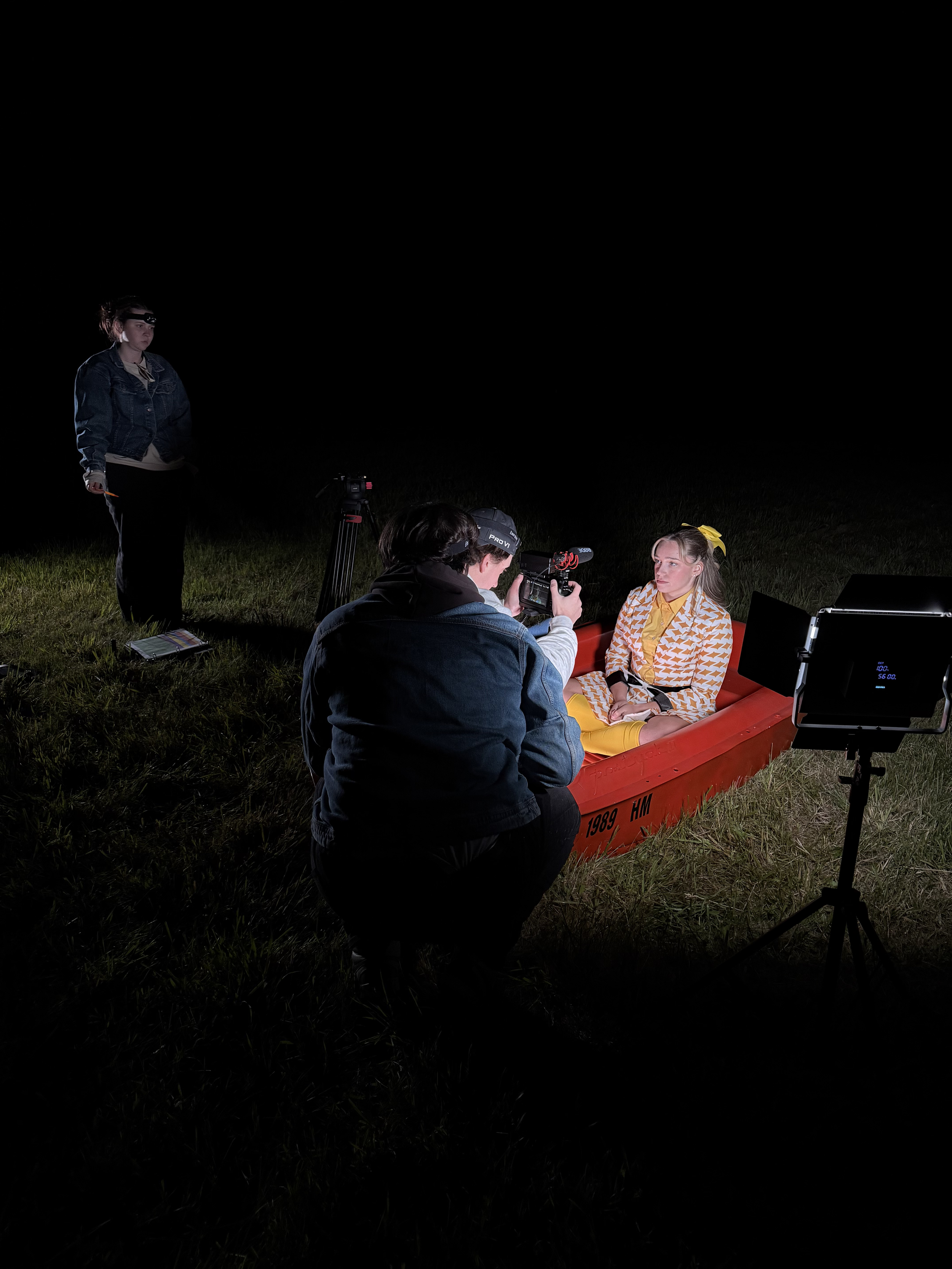 A woman in vintage clothing sitting in a red boat during a nighttime film shoot outside, with a crew filming her and a person standing nearby.