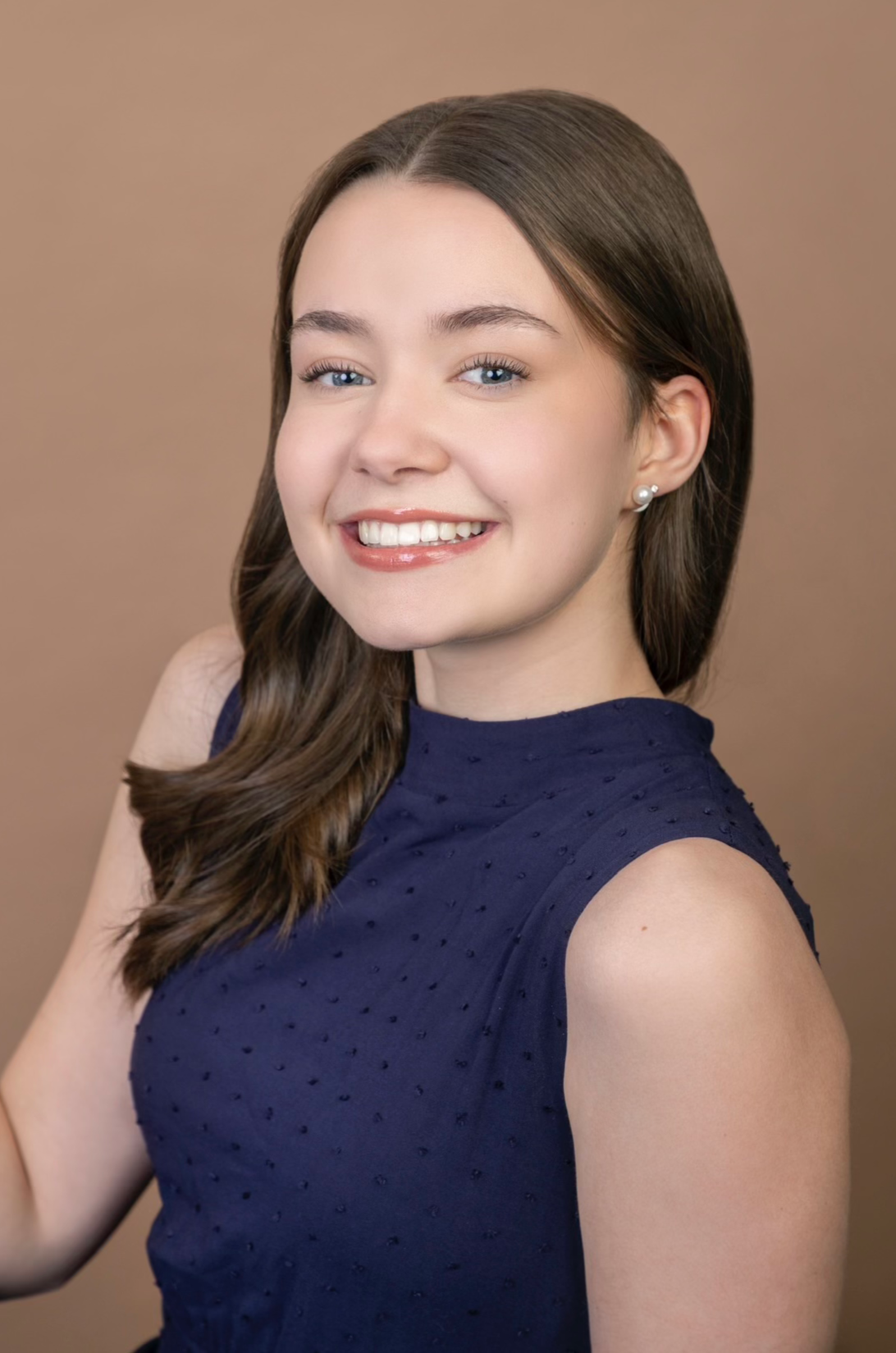 Head and shoulders portrait of a young woman with light skin, long brown hair, blue eyes, wearing pearl earrings and a navy sleeveless top with textured dots, smiling against a neutral brown background.