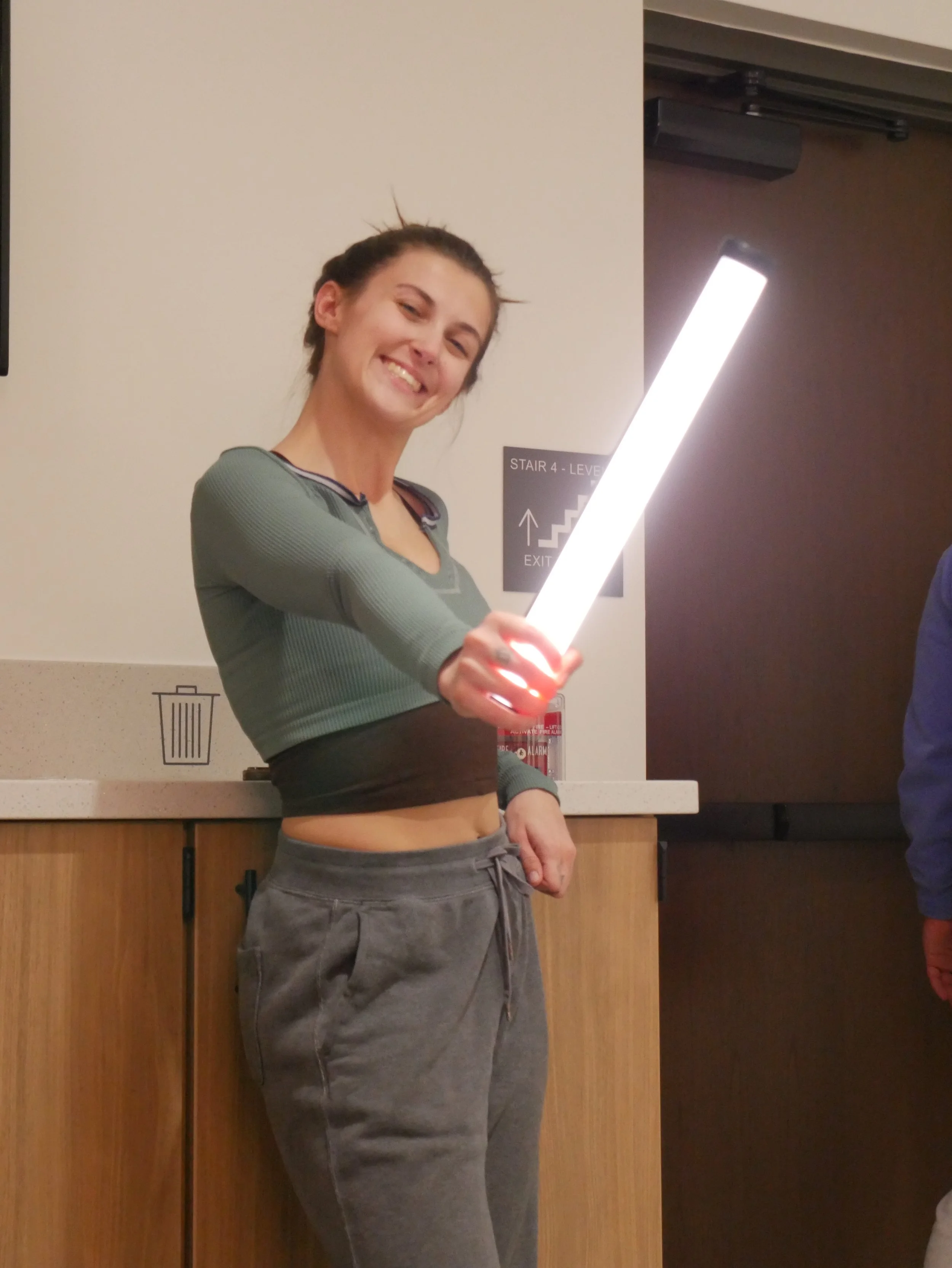 A young woman with dark hair tied back in a messy bun, smiling and holding a bright, illuminated light stick. She is wearing a cropped, long-sleeve athletic top and gray sweatpants, standing indoors near a countertop with a sign for stairs and an exit in the background.