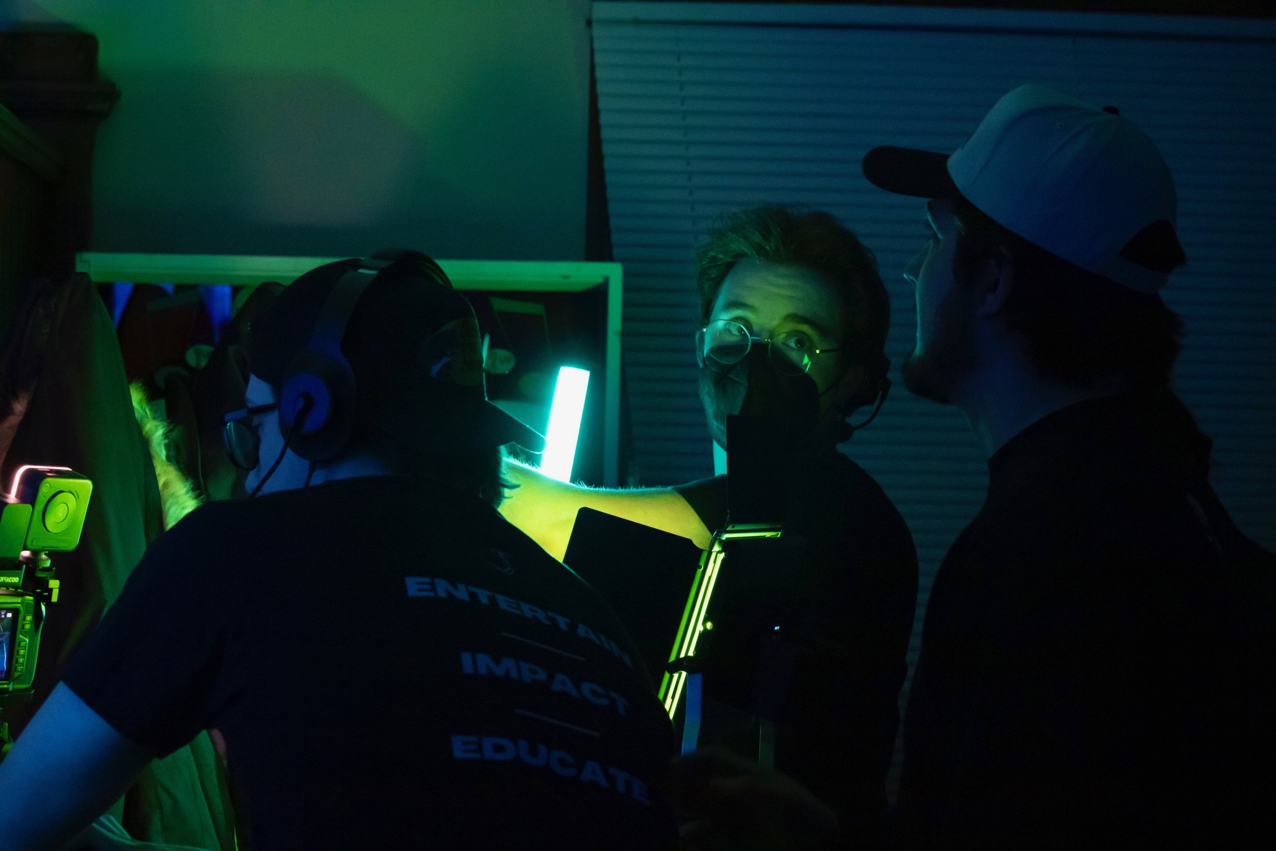 People working together in a dark room illuminated by glowing green and blue lights, wearing headphones and masks, possibly on a film or music set.