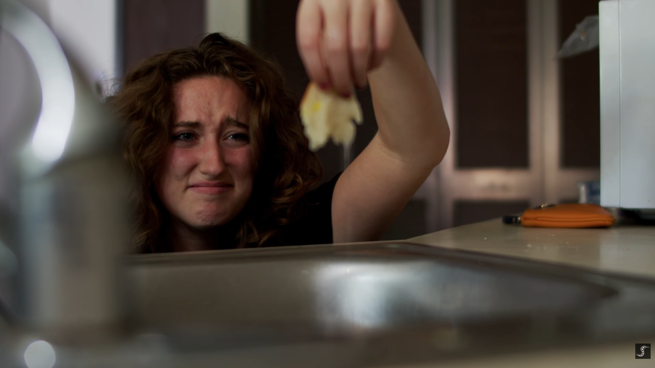 A distressed woman with curly brown hair looks upset as she reaches into a kitchen sink, holding a piece of food.