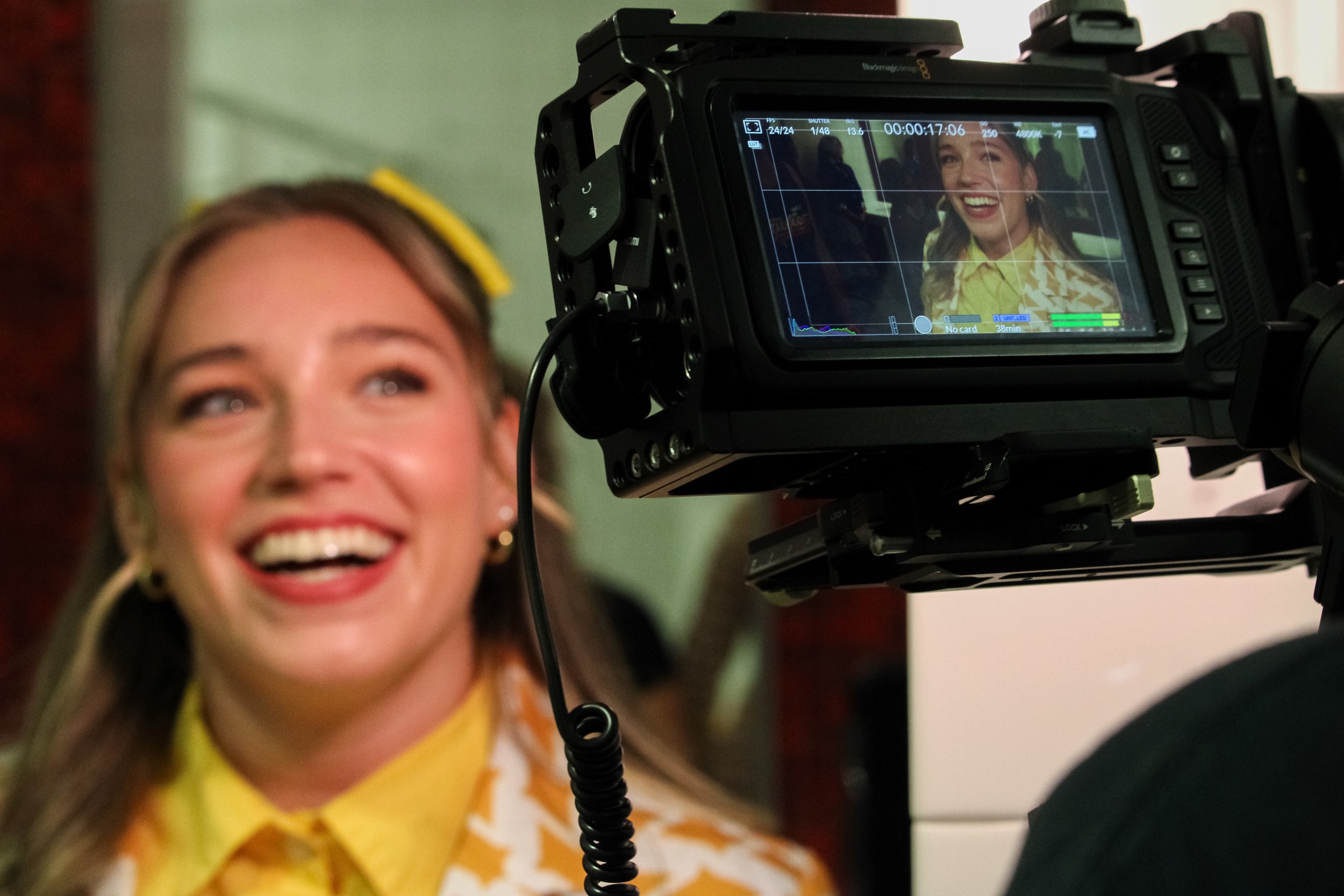 A woman smiling in front of a professional video camera, which is recording her. The camera's display shows her smiling face with a joyful expression.