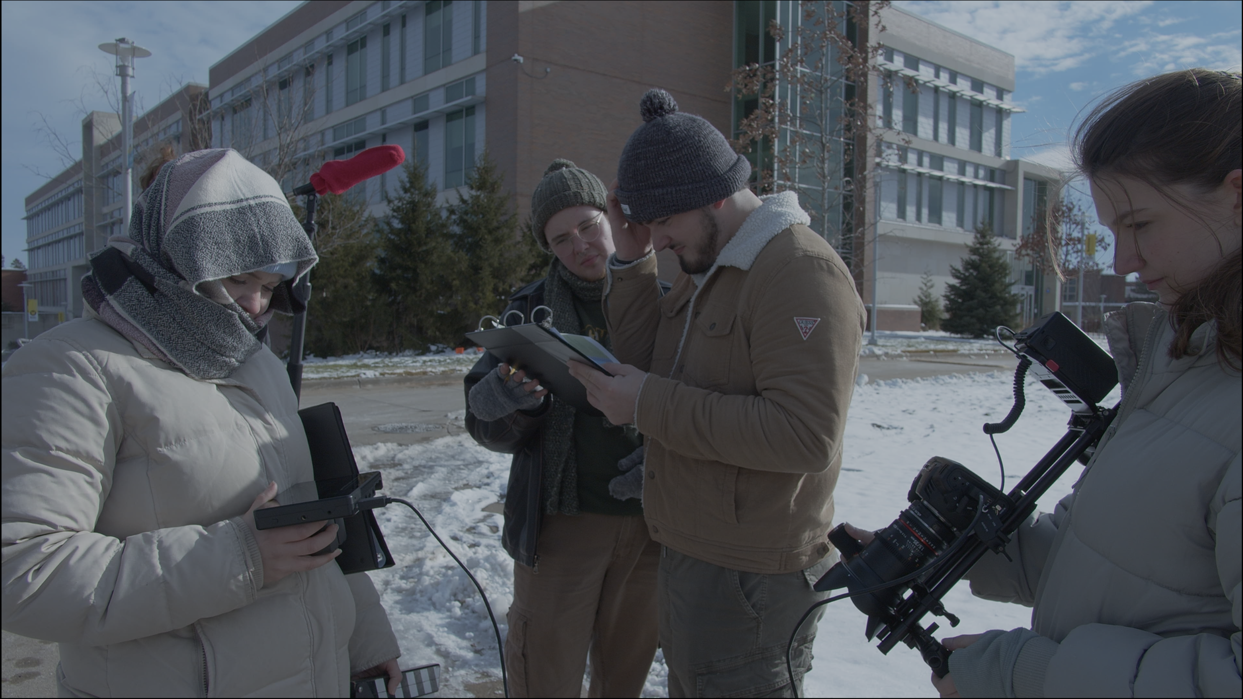 A group of four people standing outdoors on a snowy day, wearing winter clothing, engaged in a discussion while looking at equipment and documents.