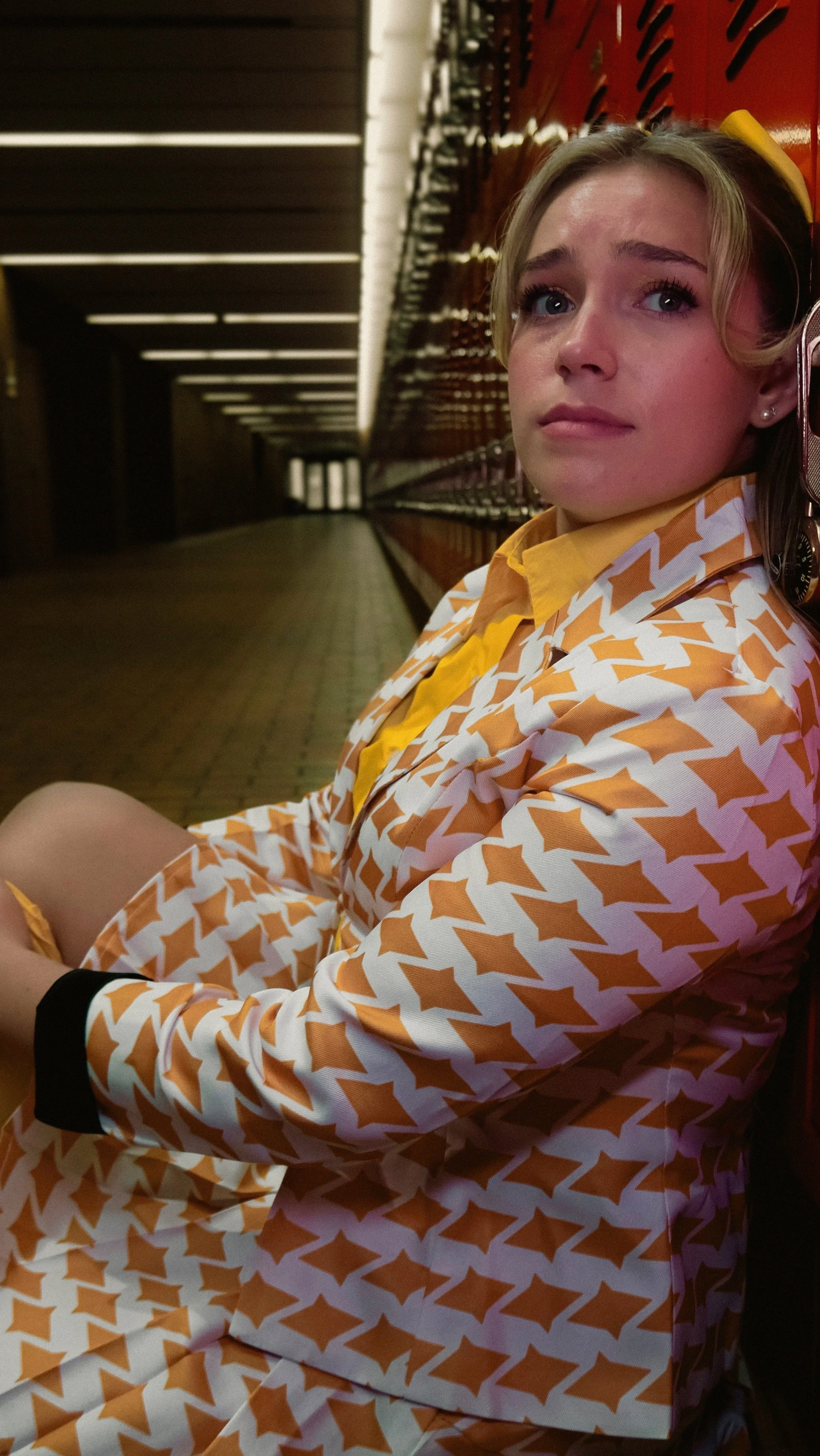 A young woman sitting on a bench, wearing a patterned orange and white outfit, with a yellow shirt underneath and a black watch. She is leaning back against a red wall with equipment storage lockers, looking upwards with a concerned expression.