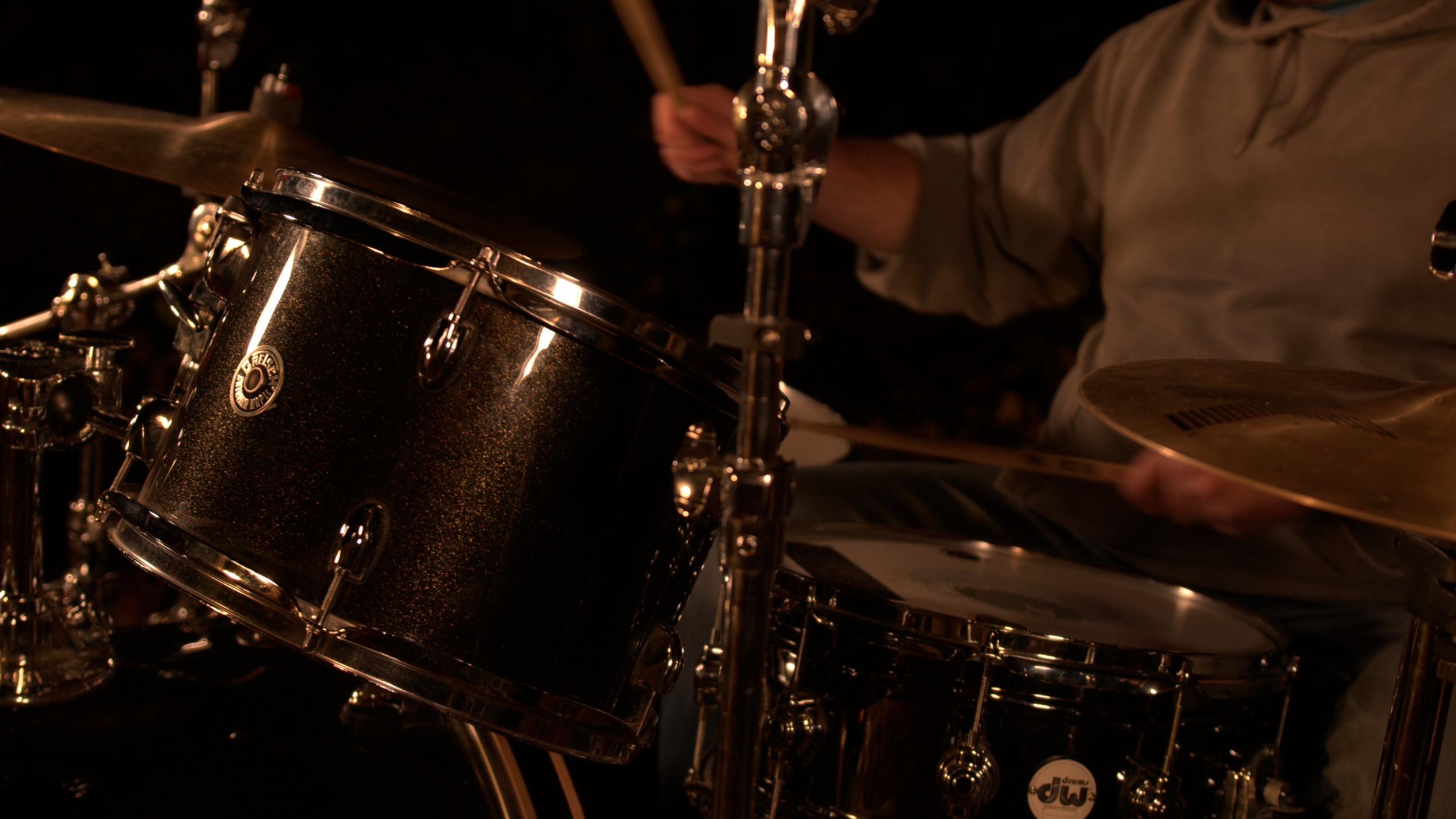 Close-up of a drum set with a black sparkle finish, featuring a snare drum and a mounted tom, with a drummer playing a cymbal in the background.