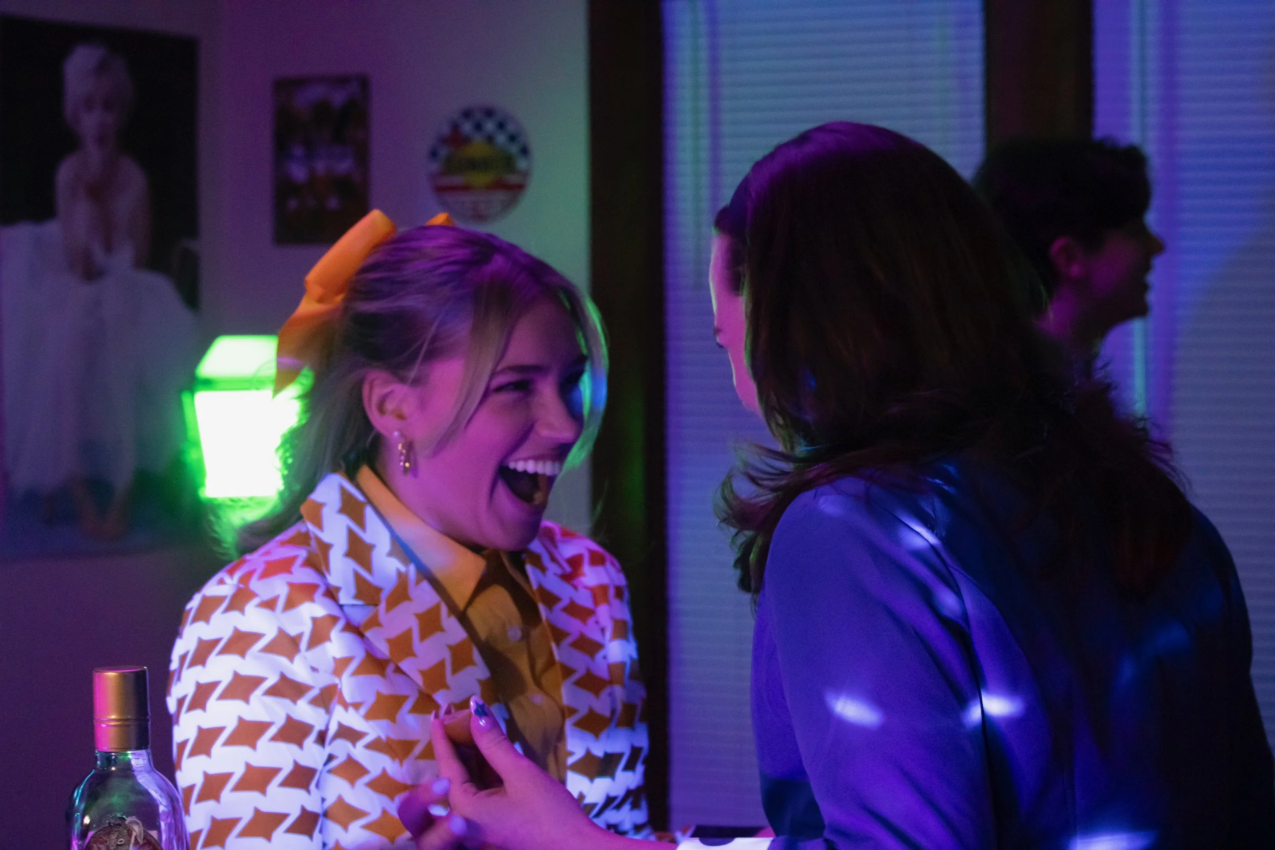 Two women are laughing and talking at a bar or nightclub, illuminated by multicolored neon lights. One woman has blonde hair with a yellow bow and is wearing a patterned jacket, while the other has dark hair and is dressed in a jacket. In the background, there are posters on the wall, including a large picture of a young girl.