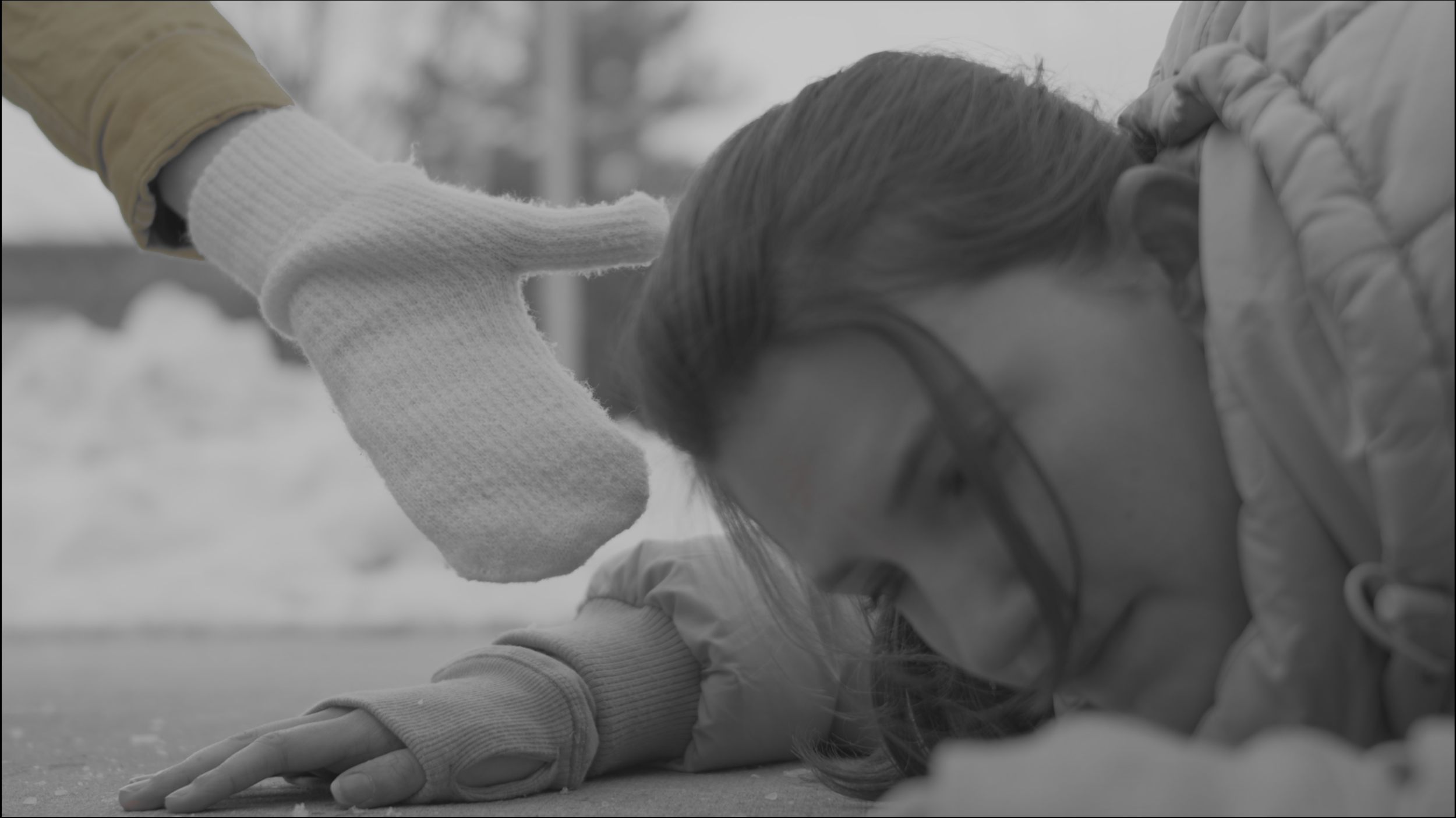 A person in a winter jacket and gloves lying on the ground with eyes closed, seemingly unconscious or injured, with another individual wearing gloves and a jacket assisting or checking on them.