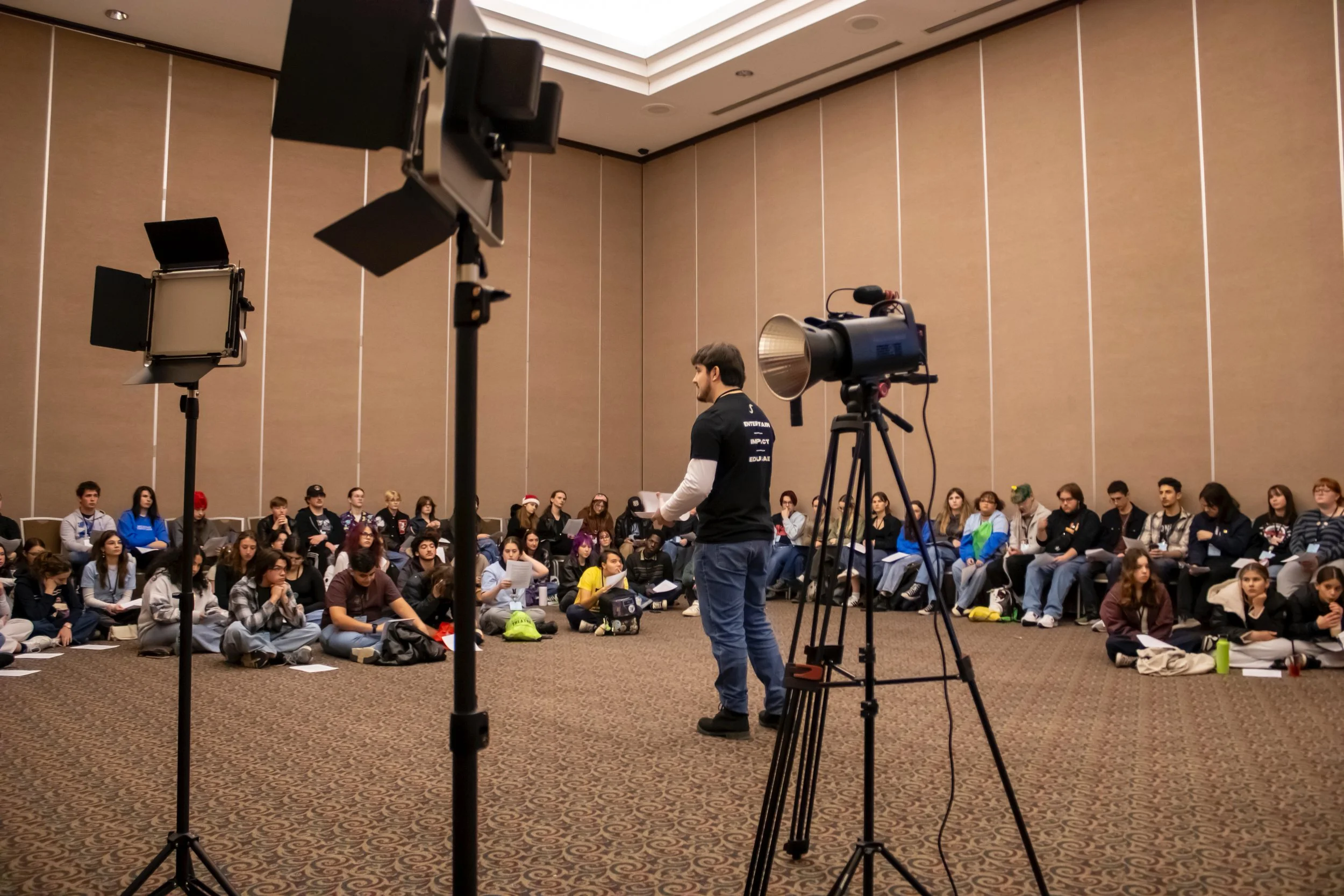 A man standing and speaking in front of an audience seated in a large conference room with beige walls, professional lighting equipment, and a camera on a tripod.
