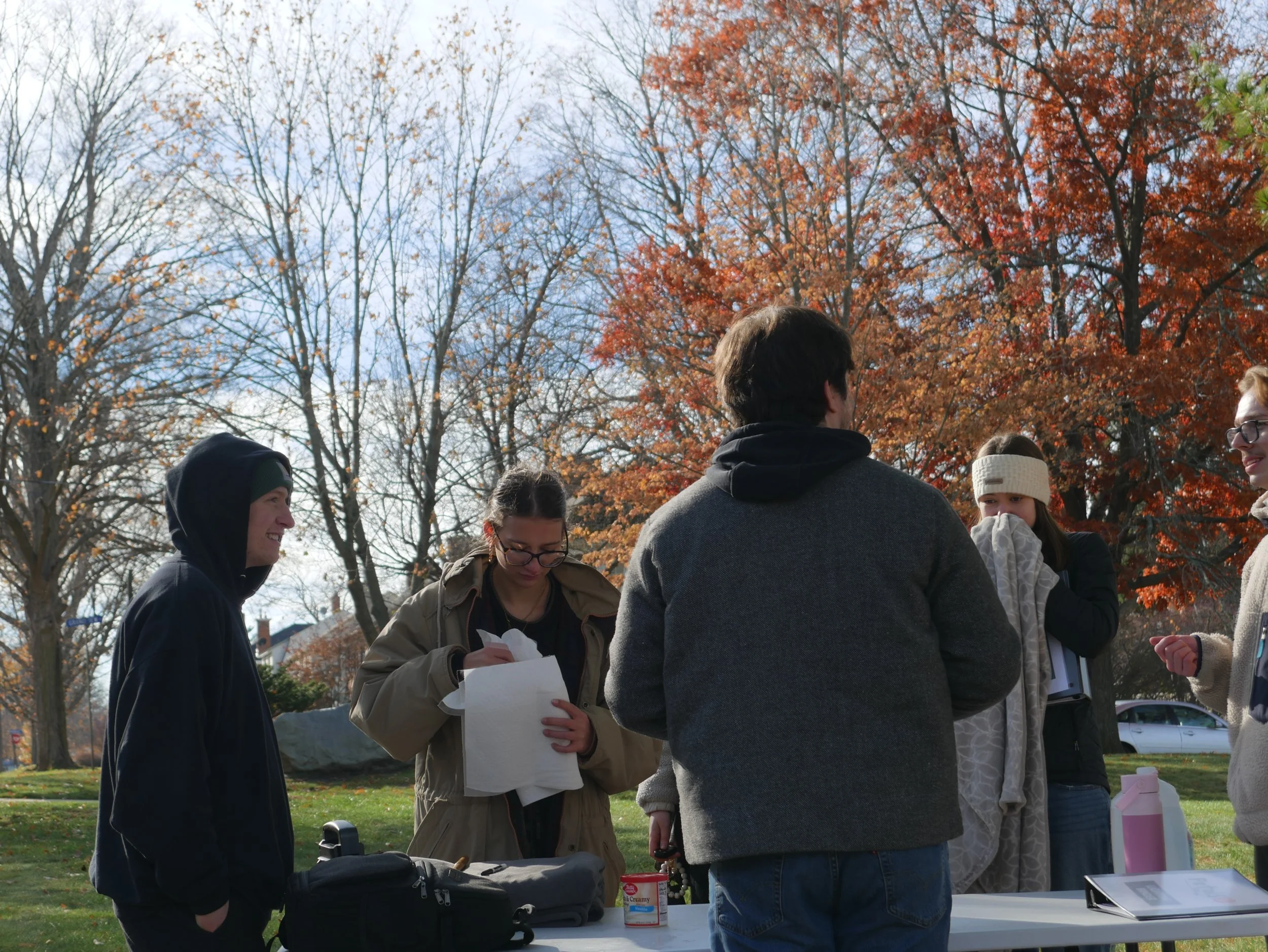 Group of five people outdoors, standing around a table, with autumn trees and a partly cloudy sky in the background.