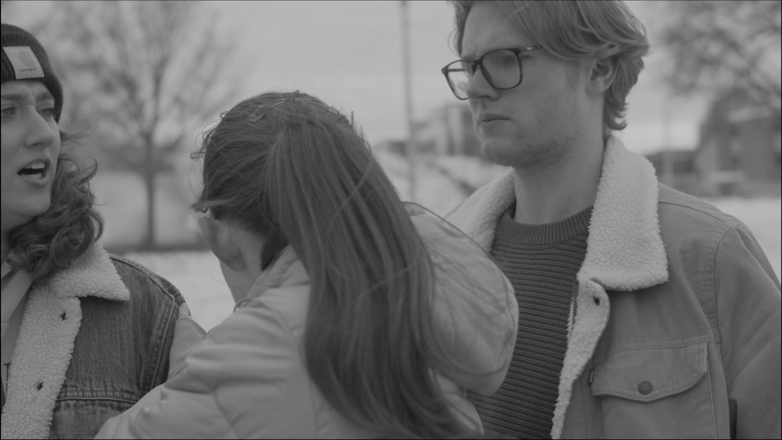 Three people outside, one woman with curly hair and a beanie speaking, and a man wearing glasses, and a girl with long hair, all engaged in a conversation during a cloudy day.