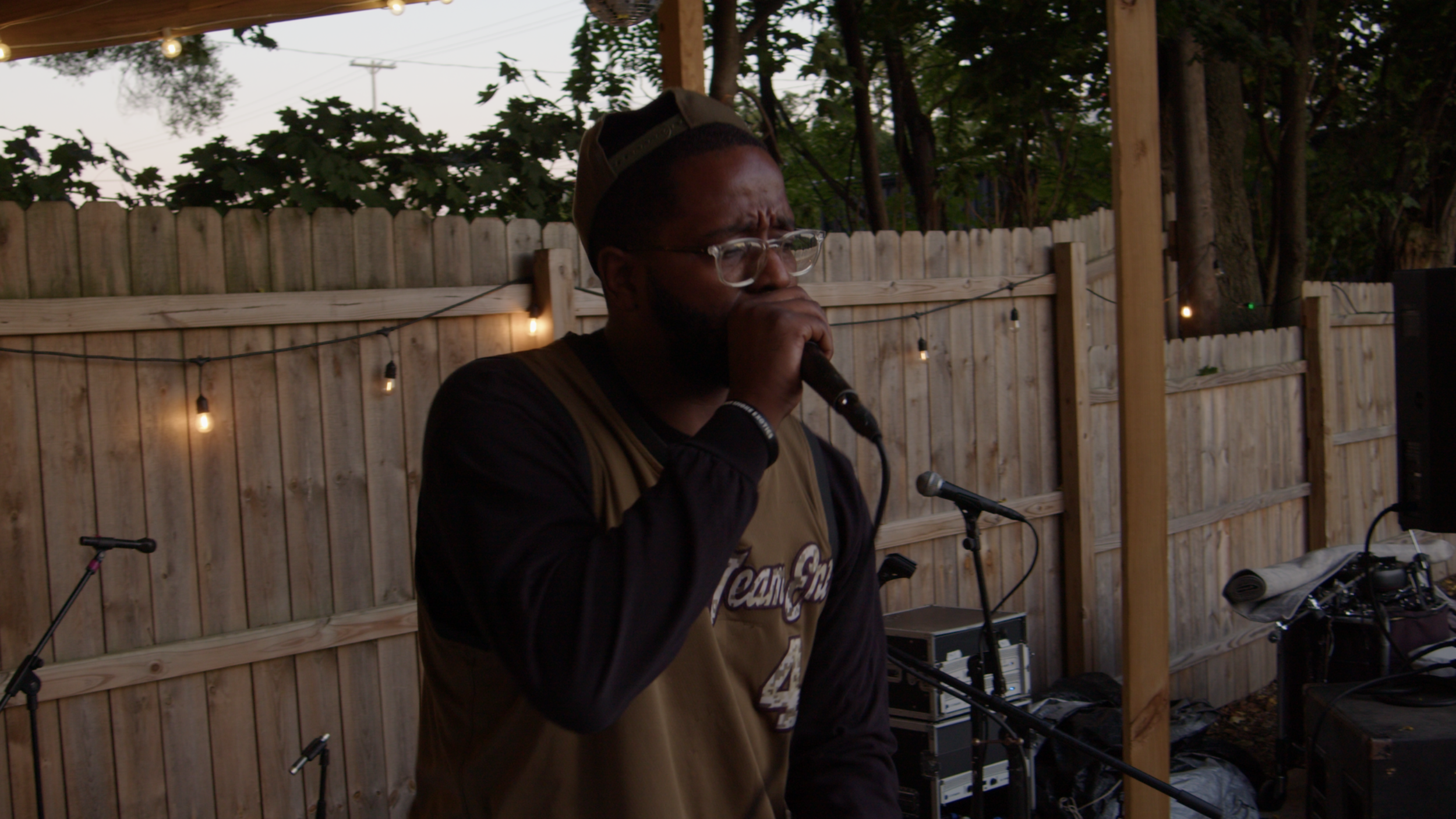 A man wearing glasses, a cap, and a brown and black sports jersey is singing or speaking into a microphone at an outdoor event, with a wooden fence and string lights in the background.