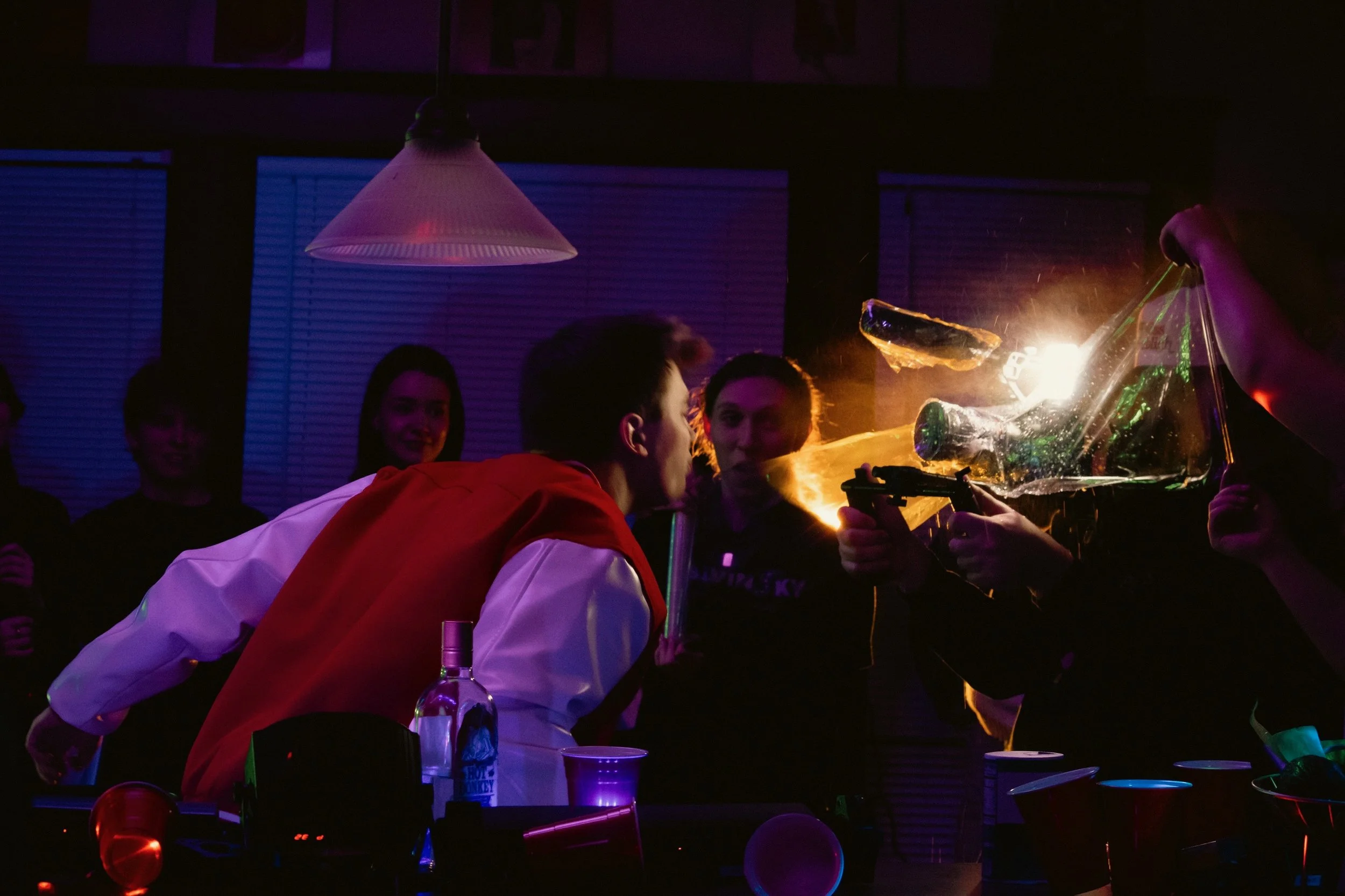 A person holding a glass bottle with flames coming out, surrounded by shadowy figures at a party with colorful lighting.