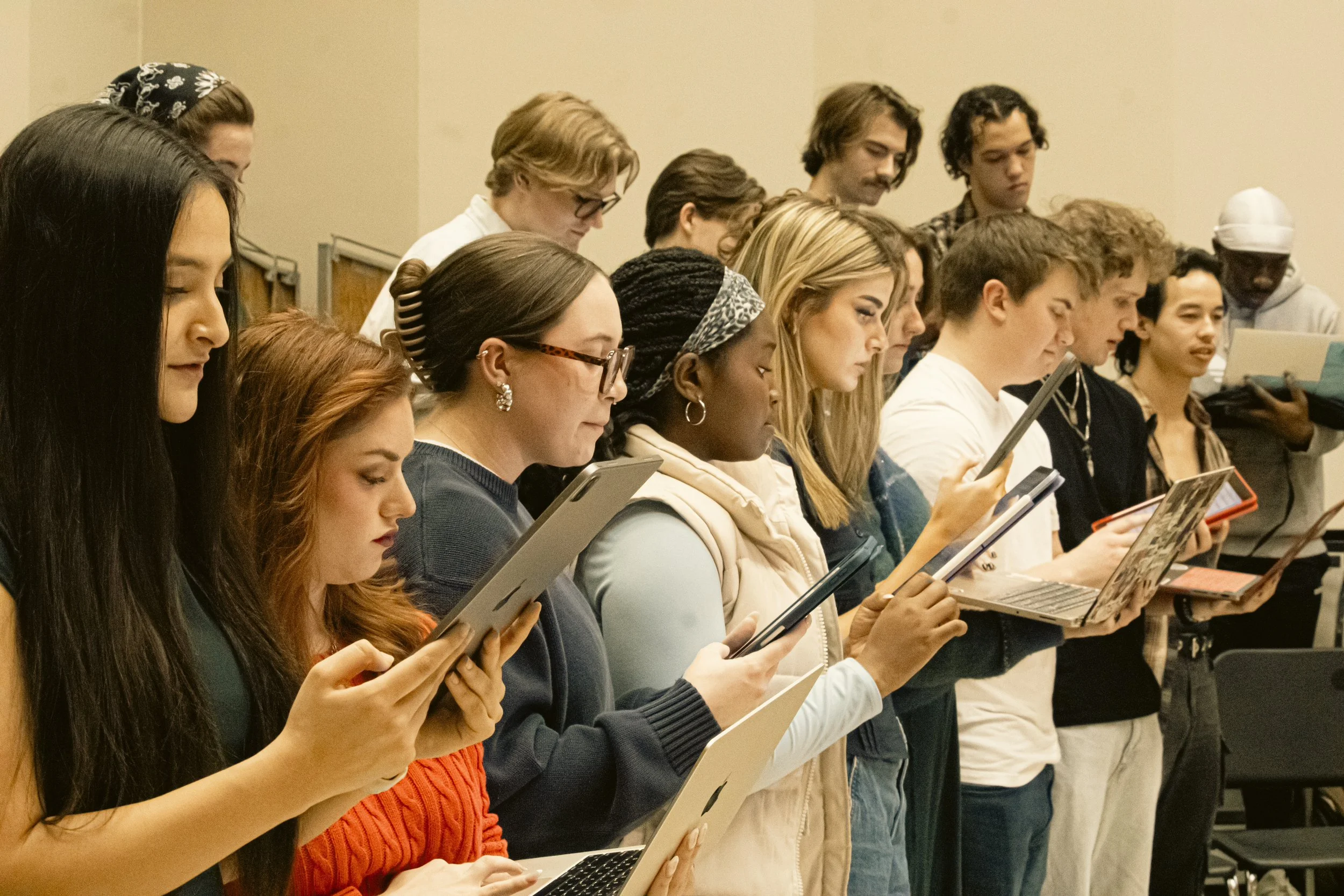 A diverse group of people, including women and men of various ethnicities, standing in a row and looking at electronic devices such as tablets, smartphones, and laptops.
