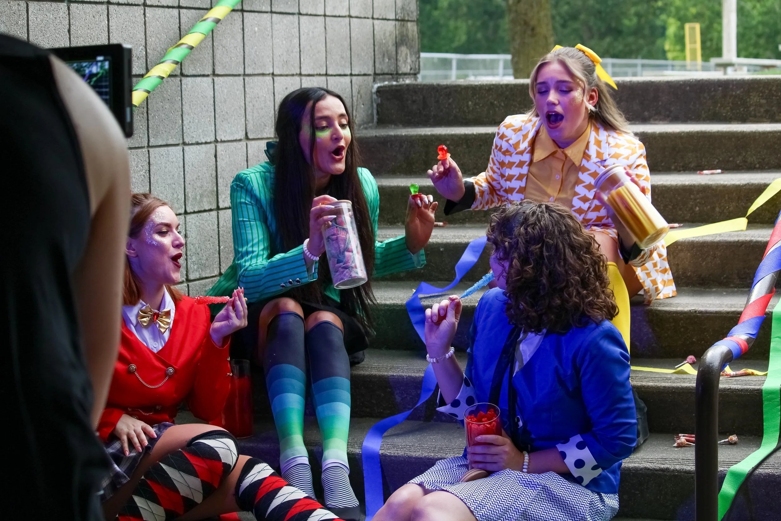 Group of women in colorful costumes sitting on outdoor steps, holding candy and drinks, celebrating with party decorations.