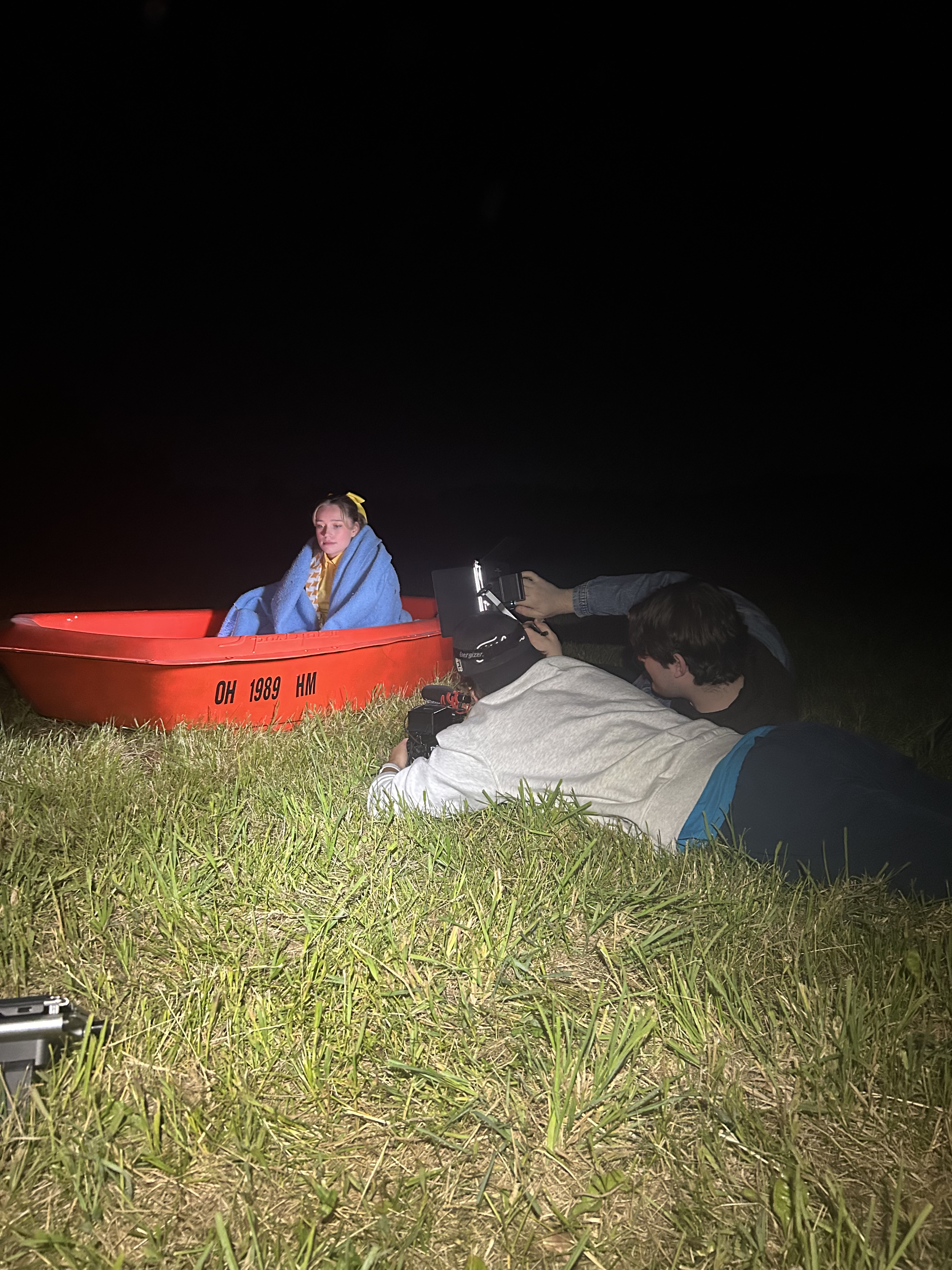 Photographers capturing a young woman wrapped in a blue blanket in a red boat at night on a grassy field.