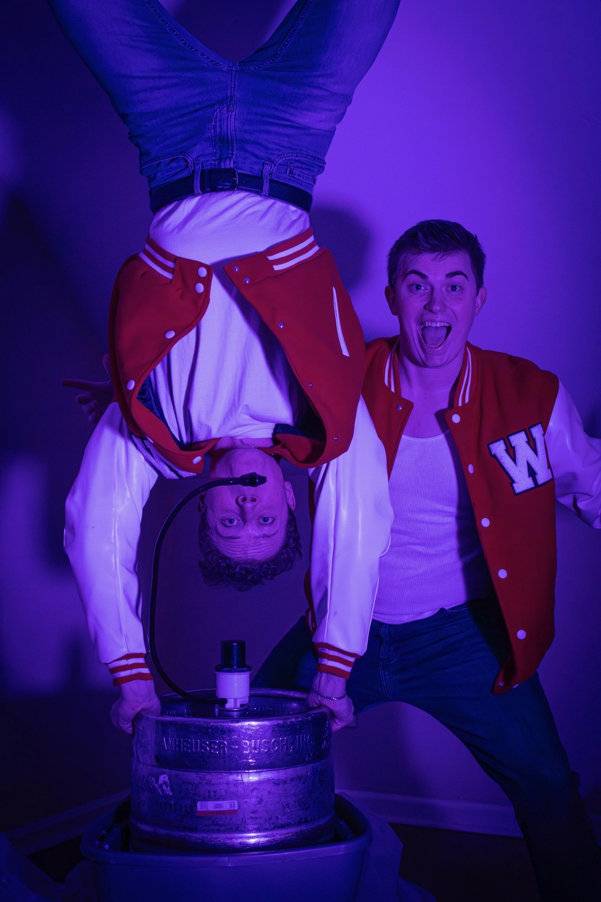 Two young men, one upside down, in a room with purple lighting, with one holding a keg. The man on the right is wearing a red and white letterman jacket with a large 'W' on it, and he is smiling at the camera. The other man is upside down, wearing a red jacket over a white shirt, and appears to be surprised or excited.