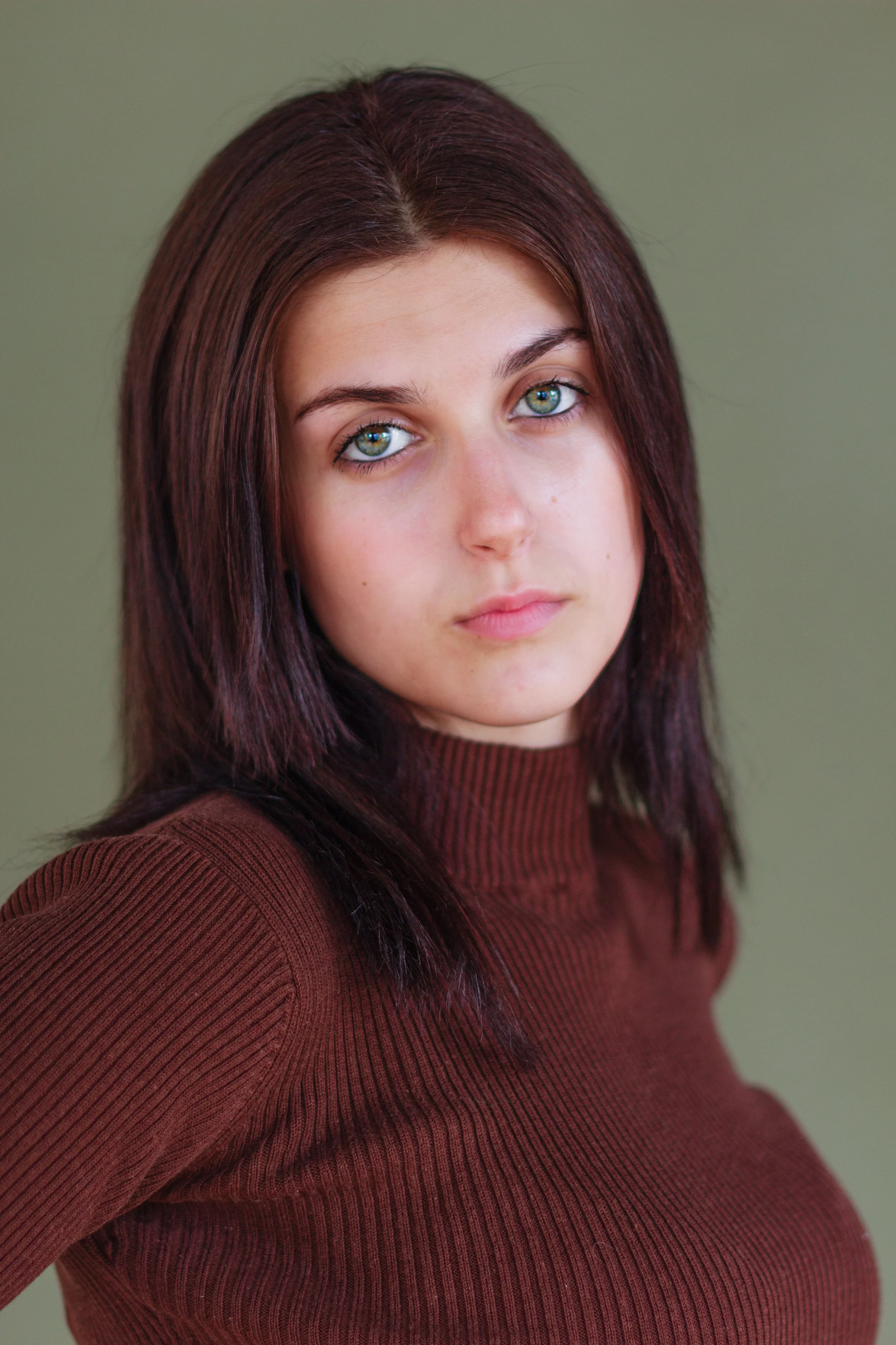 A young woman with shoulder-length dark hair wearing a brown turtleneck sweater against a plain green background.