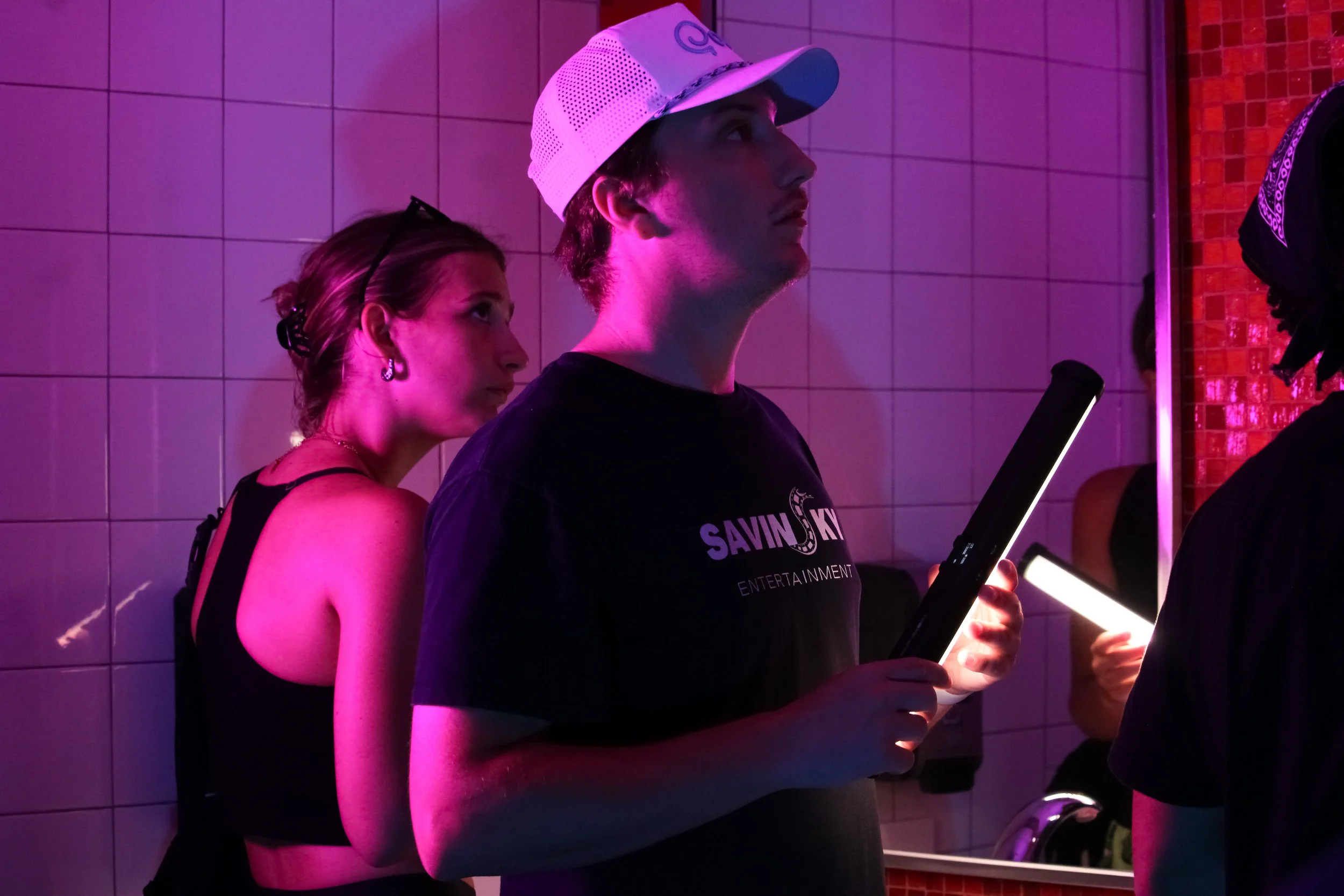 Young man holding a tablet and a light saber standing next to a woman in a black top and earrings, in a room with pink lighting and tiled walls.