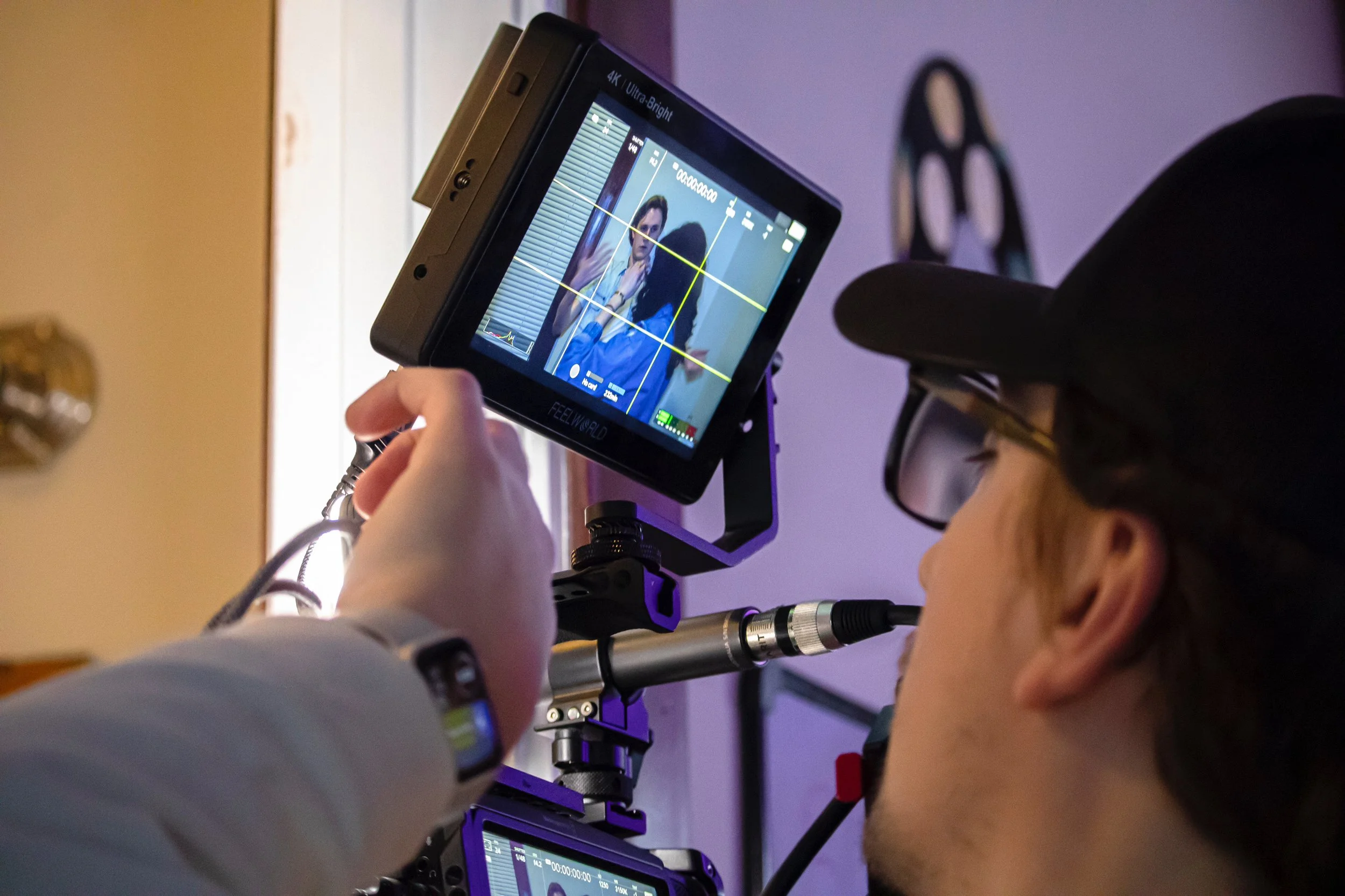 A person operating a professional camera and monitor, filming a woman in a yellow jacket and blue outfit in an indoor setting with purple lighting.