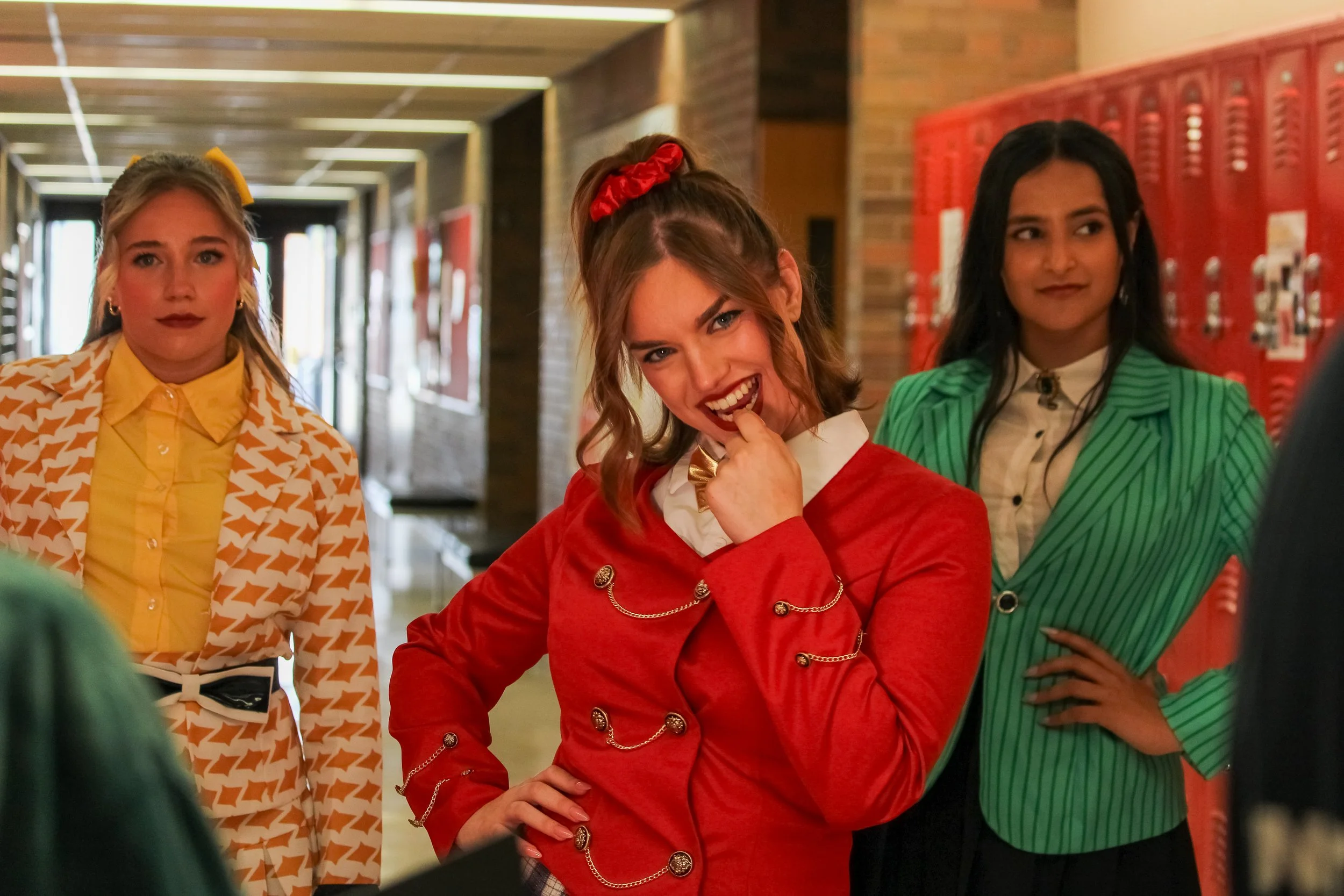 Four teenage girls standing in a school hallway with red lockers. The girl in the middle is smiling and biting her finger, wearing a red blazer and white shirt. The girl on the left wears a yellow shirt with a patterned jacket, and the girl on the right wears a green striped blazer with a white shirt.