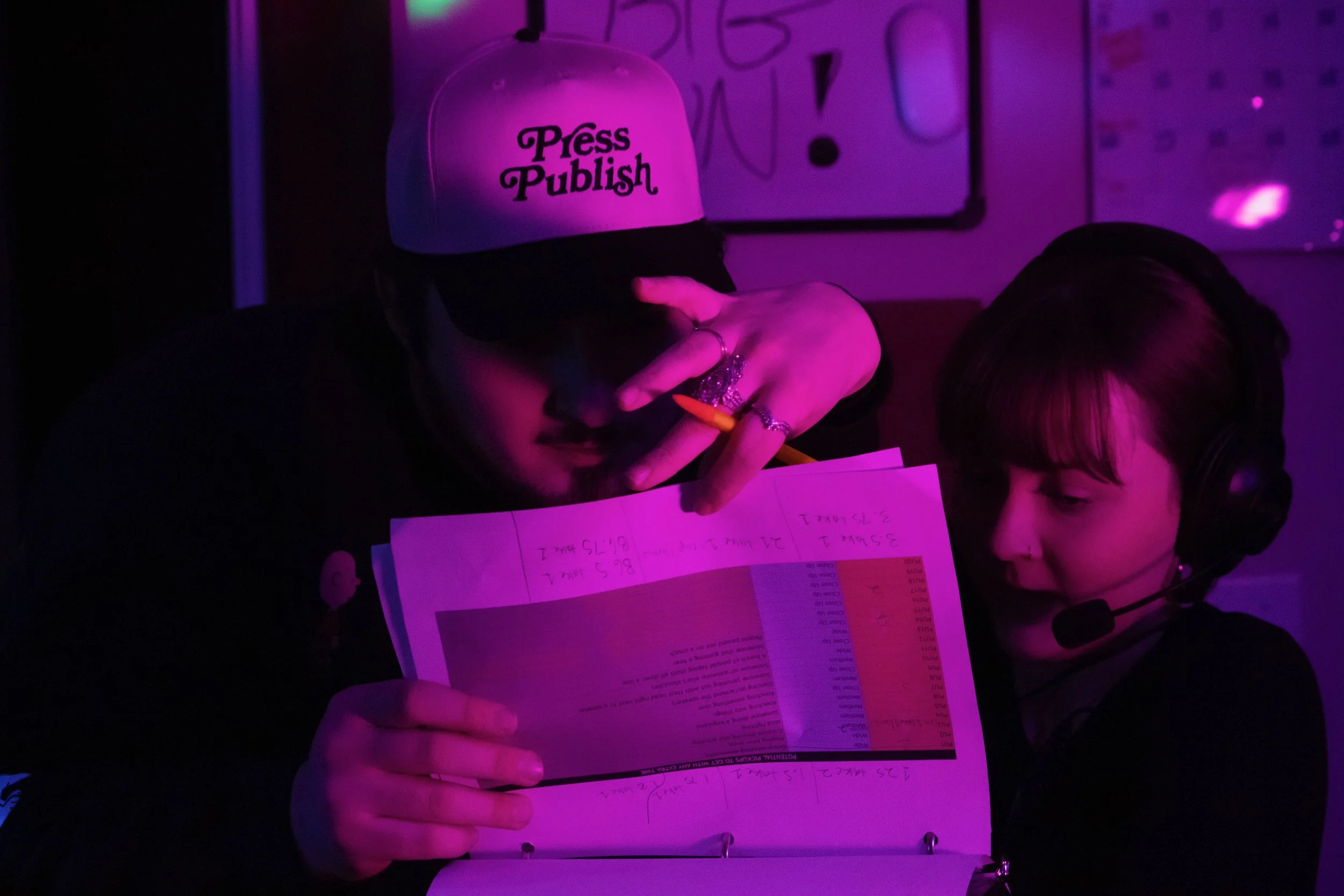 Two people working together under purple lighting. The person on the left is wearing a white cap with the words "Press Publish" and is holding papers while making a peace sign gesture. The person on the right is wearing a headset and looking at the papers.