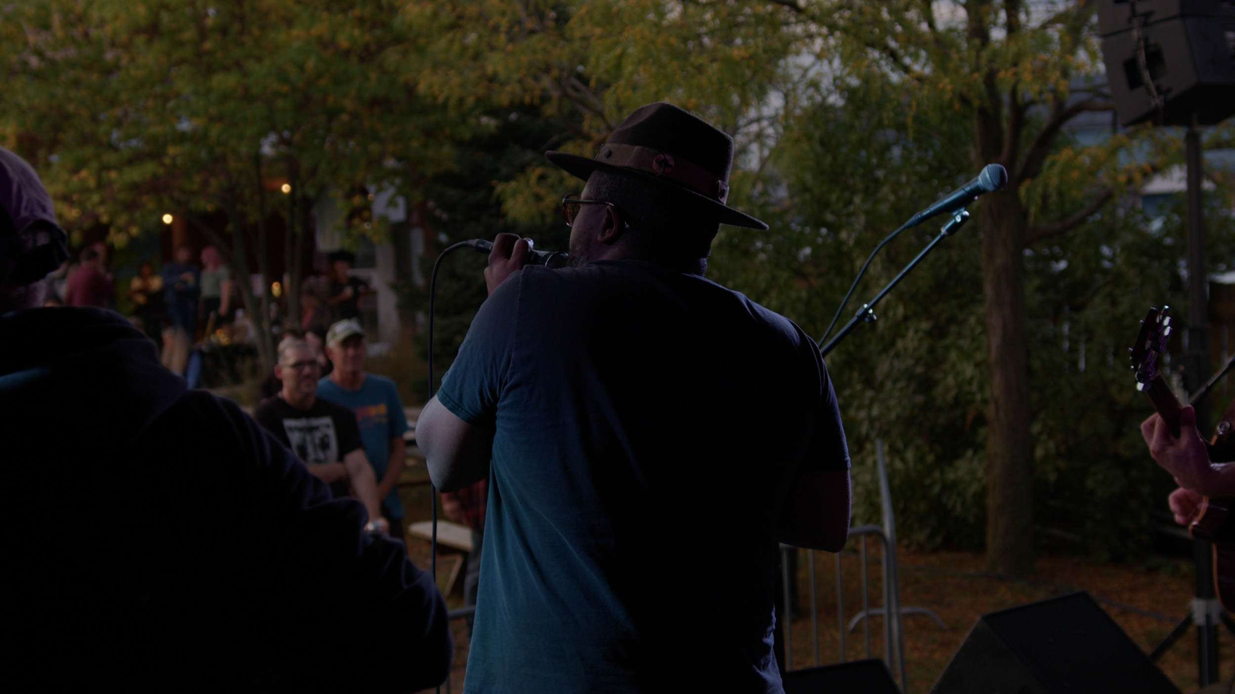 A person wearing a dark hat and blue T-shirt is holding a microphone and performing on an outdoor stage during dusk. There are people and trees in the background.