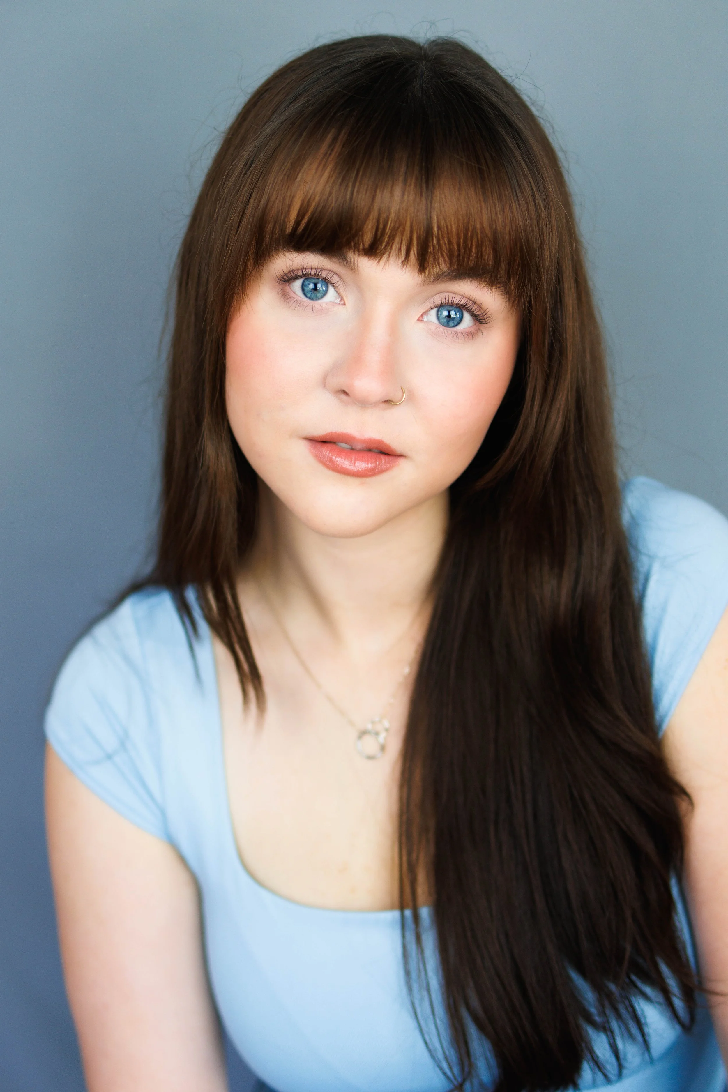A young woman with long brown hair, blue eyes, wearing a light blue top and a necklace, looking directly at the camera against a gray background.